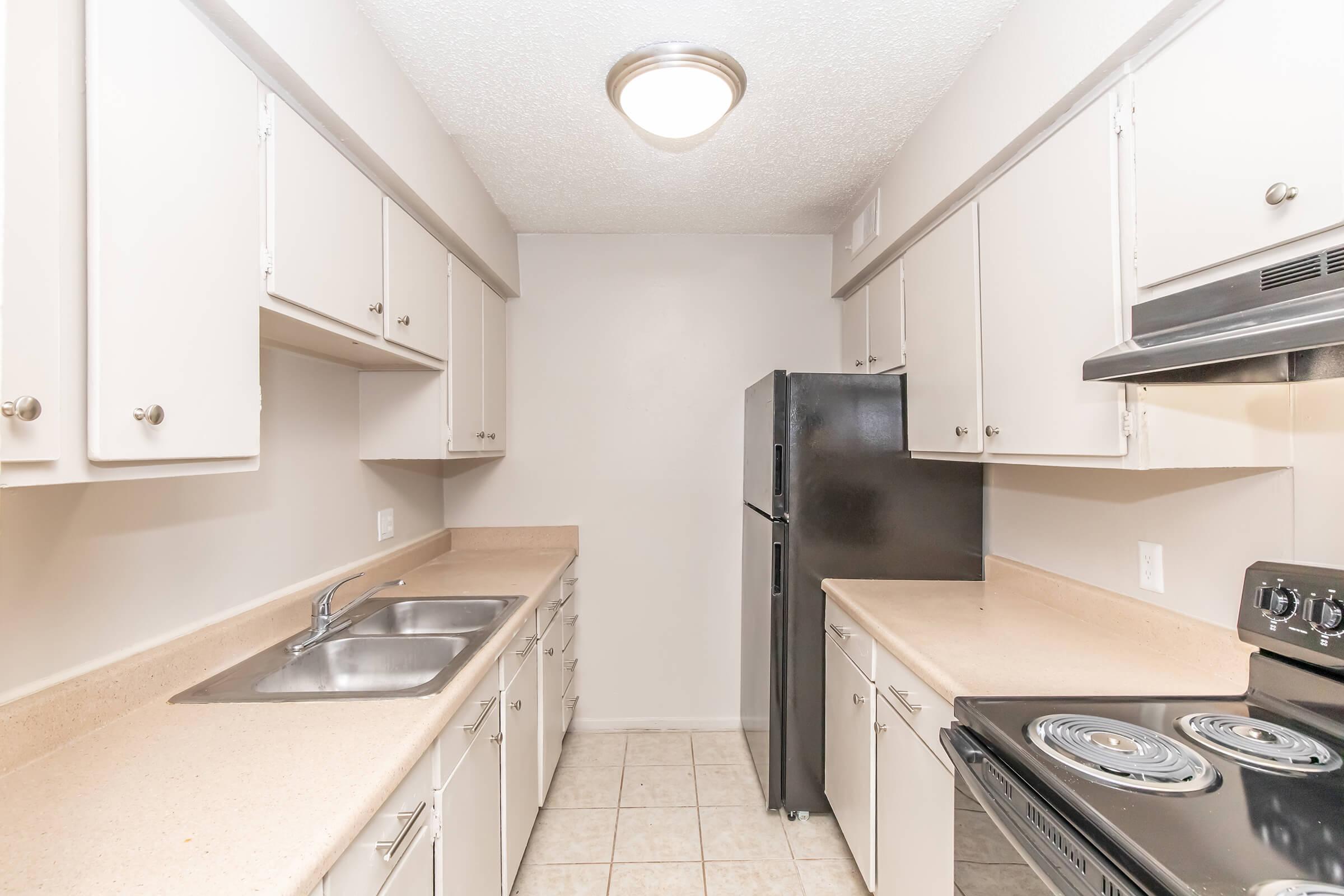 A small kitchen with light-colored cabinets, a double sink, a black refrigerator, and an electric stove. The countertops are beige, and the walls are painted in a light neutral tone. The kitchen is well-lit with overhead lighting and features tile flooring.