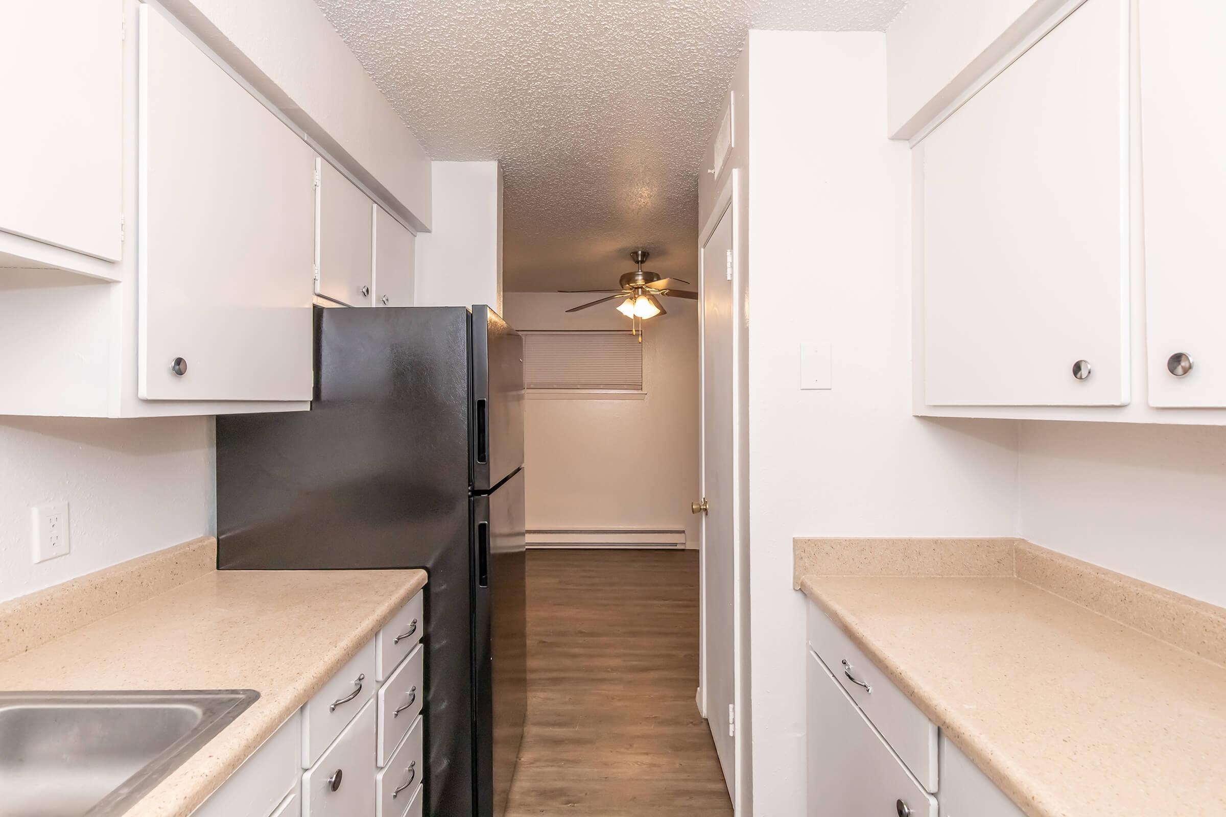 View of a modern kitchen featuring white cabinets, a black refrigerator, and beige countertops. The kitchen leads to a well-lit living area, with laminate flooring and a ceiling fan visible in the background. The space appears clean and spacious, ideal for cooking and entertaining.