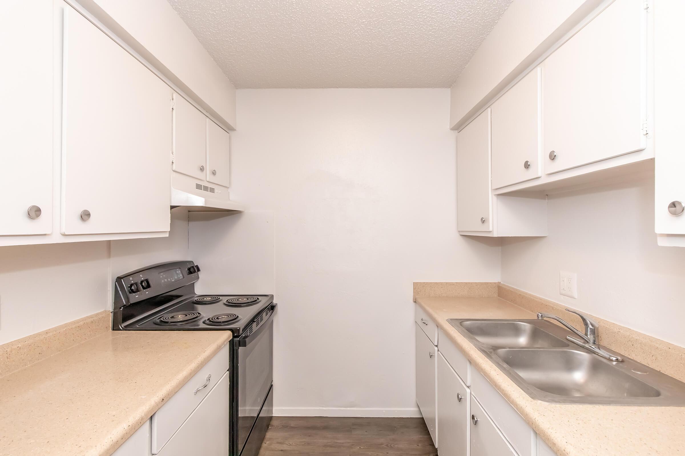 A modern kitchen featuring white cabinetry, a double sink, and a stainless steel stove with an oven. The countertops are light-colored, and the walls are painted white, creating a clean and spacious atmosphere. The flooring is a dark wood laminate.