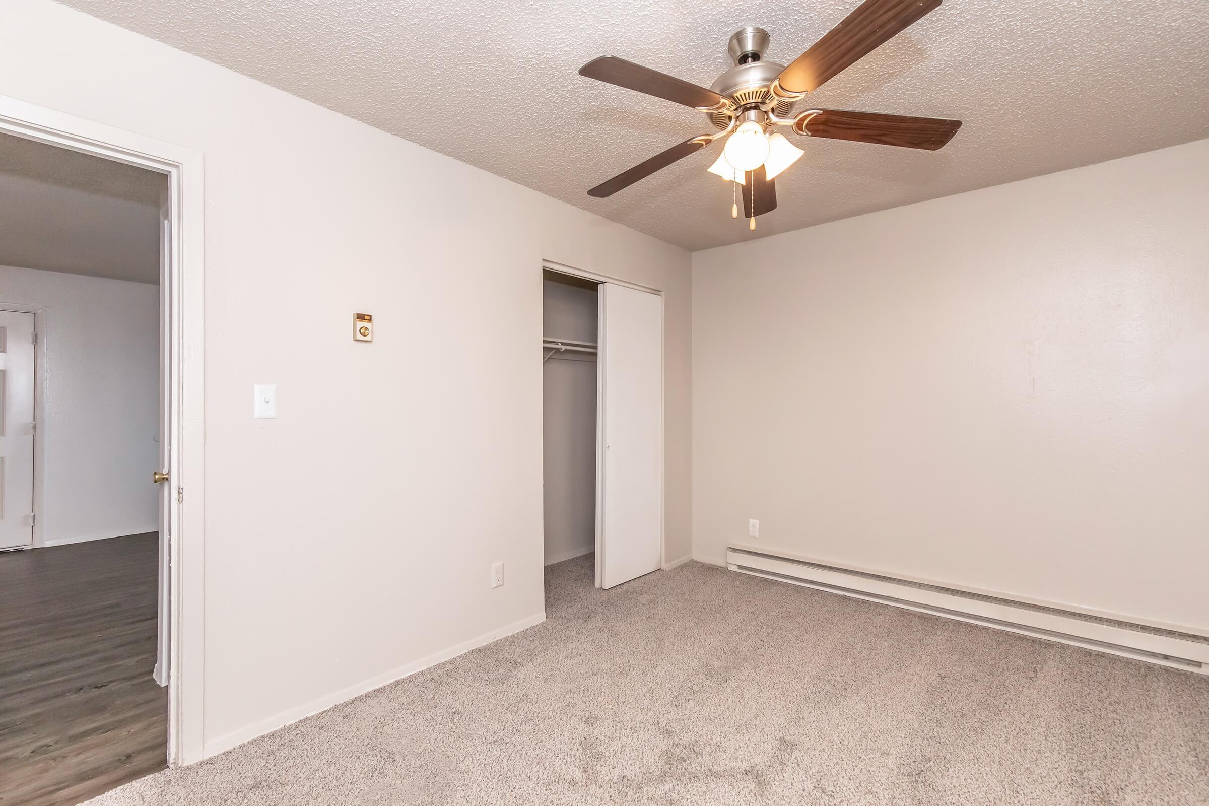 A spacious room featuring light beige walls, a ceiling fan with wooden blades, and plush carpet flooring. To the left, there is a door leading to another area, and opposite is a closet with sliding doors. The room is well-lit, creating a warm and inviting atmosphere.