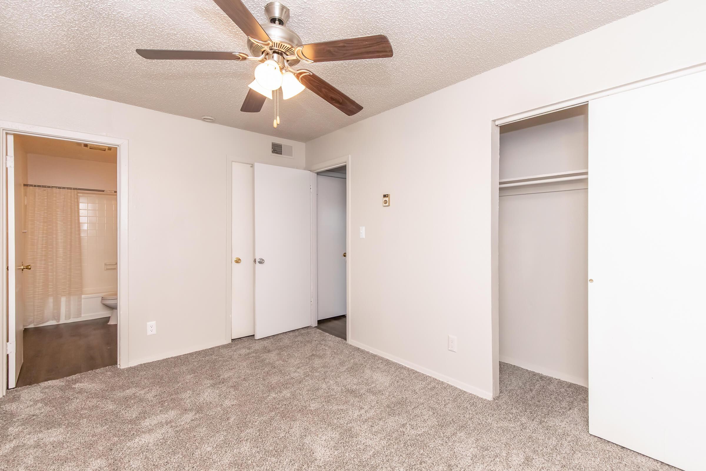 A well-lit bedroom with a ceiling fan, light-colored walls, and plush carpet. There are two closets with white doors, one slightly ajar. In the background, a bathroom is partially visible through a door, indicating the layout of the space. Overall, the room appears clean and inviting.