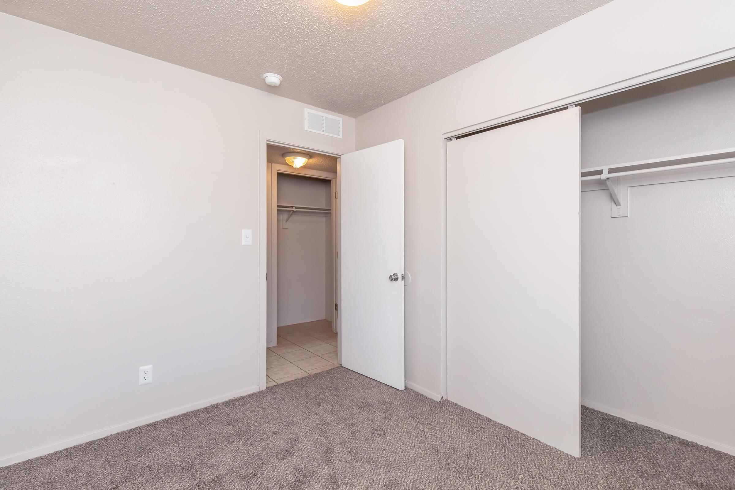 A view of a small, empty bedroom featuring light-colored walls and carpet. The room includes a closet with sliding doors and a door leading to a small, empty storage area. A ceiling light fixture illuminates the space, which is unfurnished and ready for personal decoration.