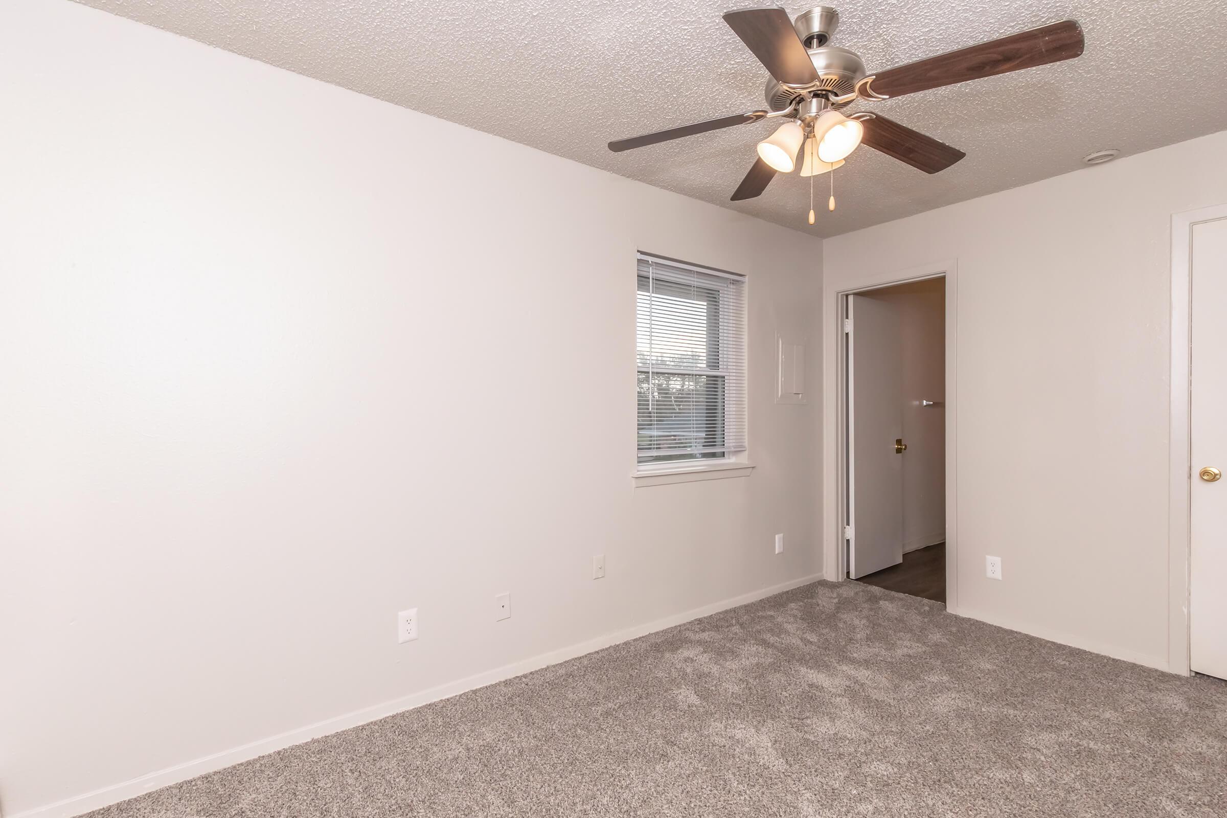 A sparse room featuring light gray walls, a window with blinds, and a ceiling fan with light fixtures. The floor is covered in plush gray carpet, and there is a door leading to another space. The room appears bright and freshly painted, offering a clean and neutral aesthetic.