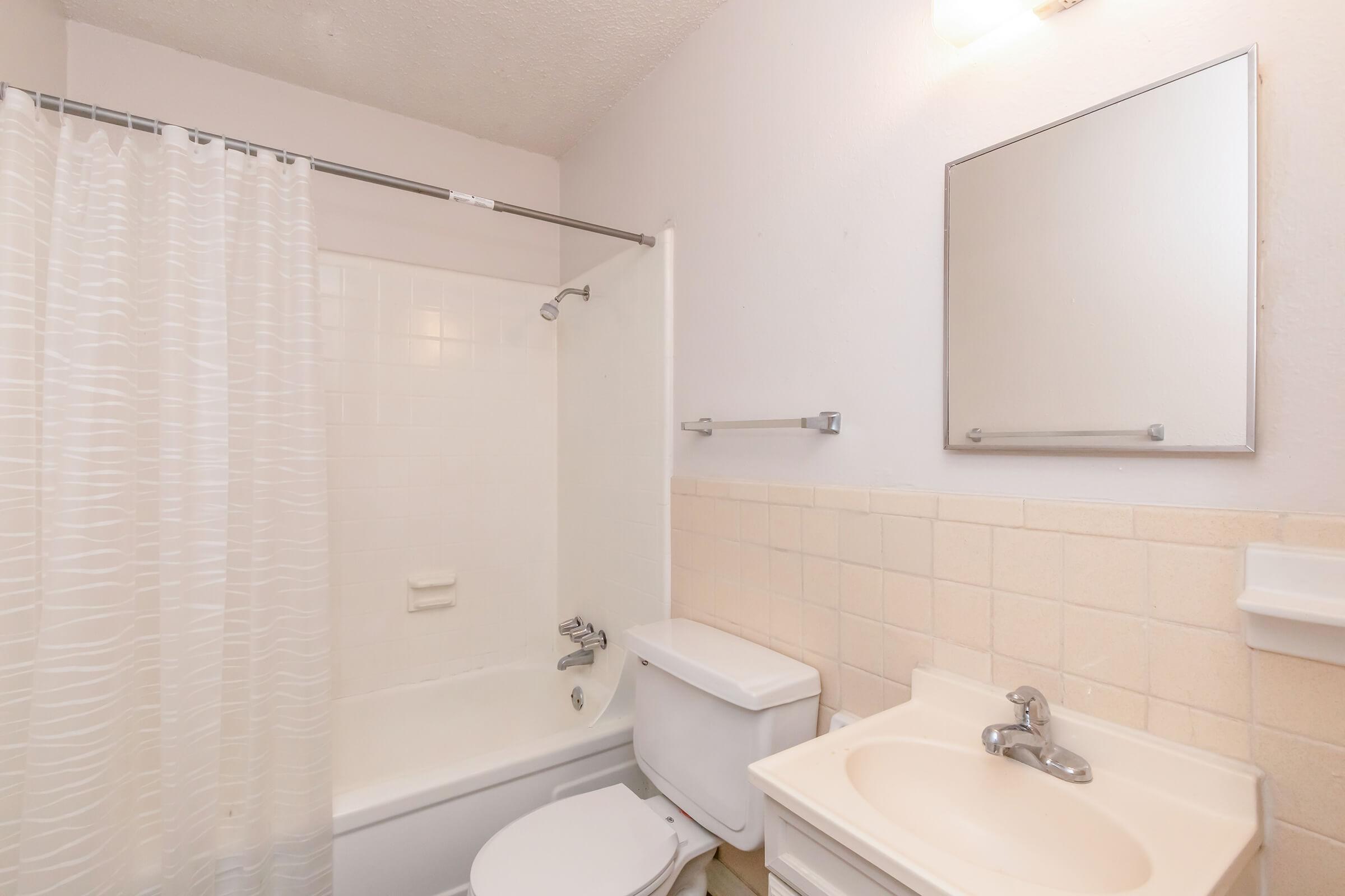 A clean, small bathroom featuring a white bathtub with a shower curtain, a toilet, a sink with a countertop, and a mirror above the sink. The walls are painted light colors, and the floor is visible, creating a simple and functional space.