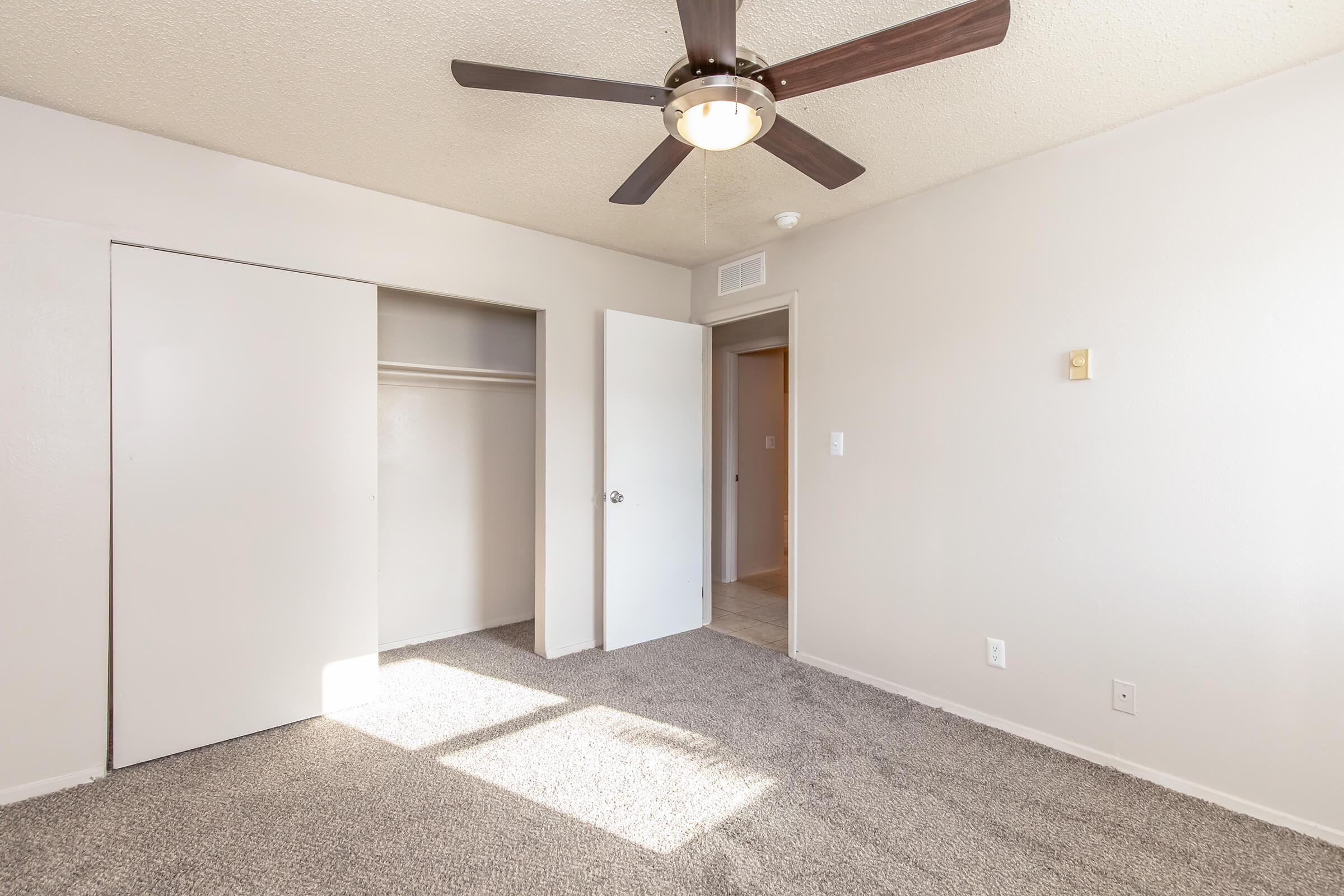 A well-lit bedroom featuring neutral-colored walls, a ceiling fan, and carpeted flooring. The room includes a closed closet with sliding doors and an open door leading to another space. Sunlight casts shadows on the floor, creating a warm atmosphere.