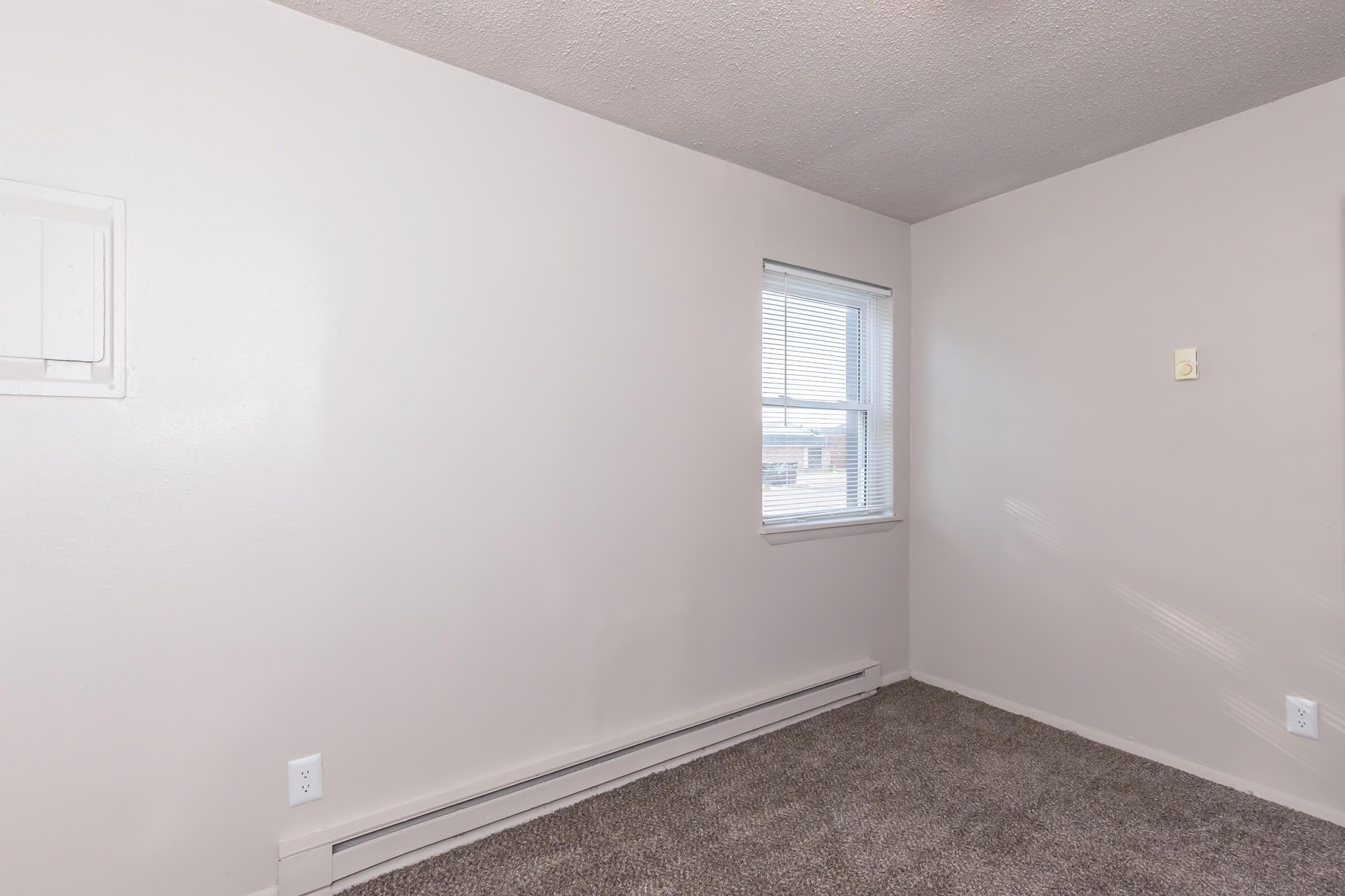 A small, empty room with light beige walls and grey carpet. It features a window with blinds letting in natural light, and there's a small wall-mounted electrical panel. The space appears clean and unfurnished, ideal for personal customization.