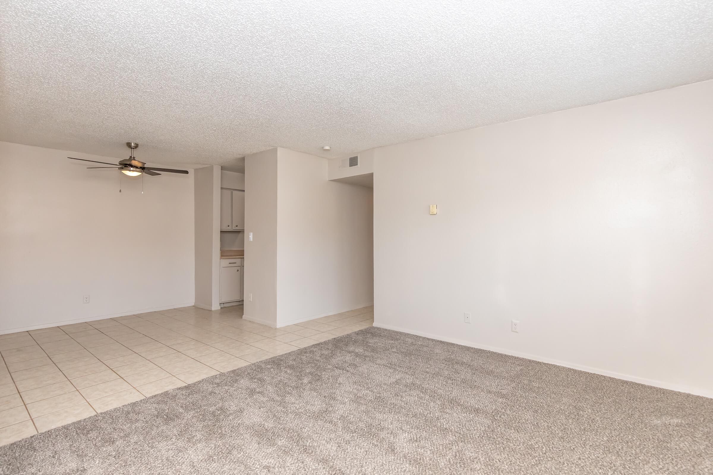 A spacious empty living area featuring light-colored walls, a textured ceiling, and tiled flooring. On one side, there is a ceiling fan, and a doorway leads to a kitchen area with cabinetry. The opposite side has plush carpet, creating a cozy contrast with the tiled portion.