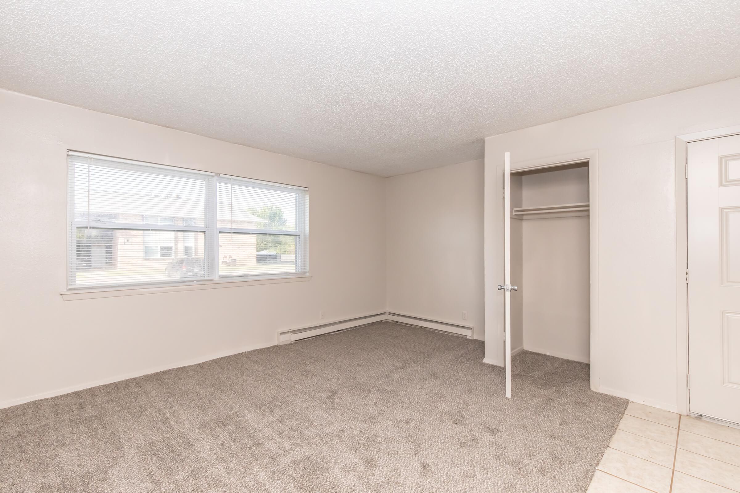 A spacious, empty room with beige carpet and light-colored walls. It features two windows allowing natural light, a small closet with a door, and a tile section on the floor near the entrance. The room appears clean and ready for furnishing.