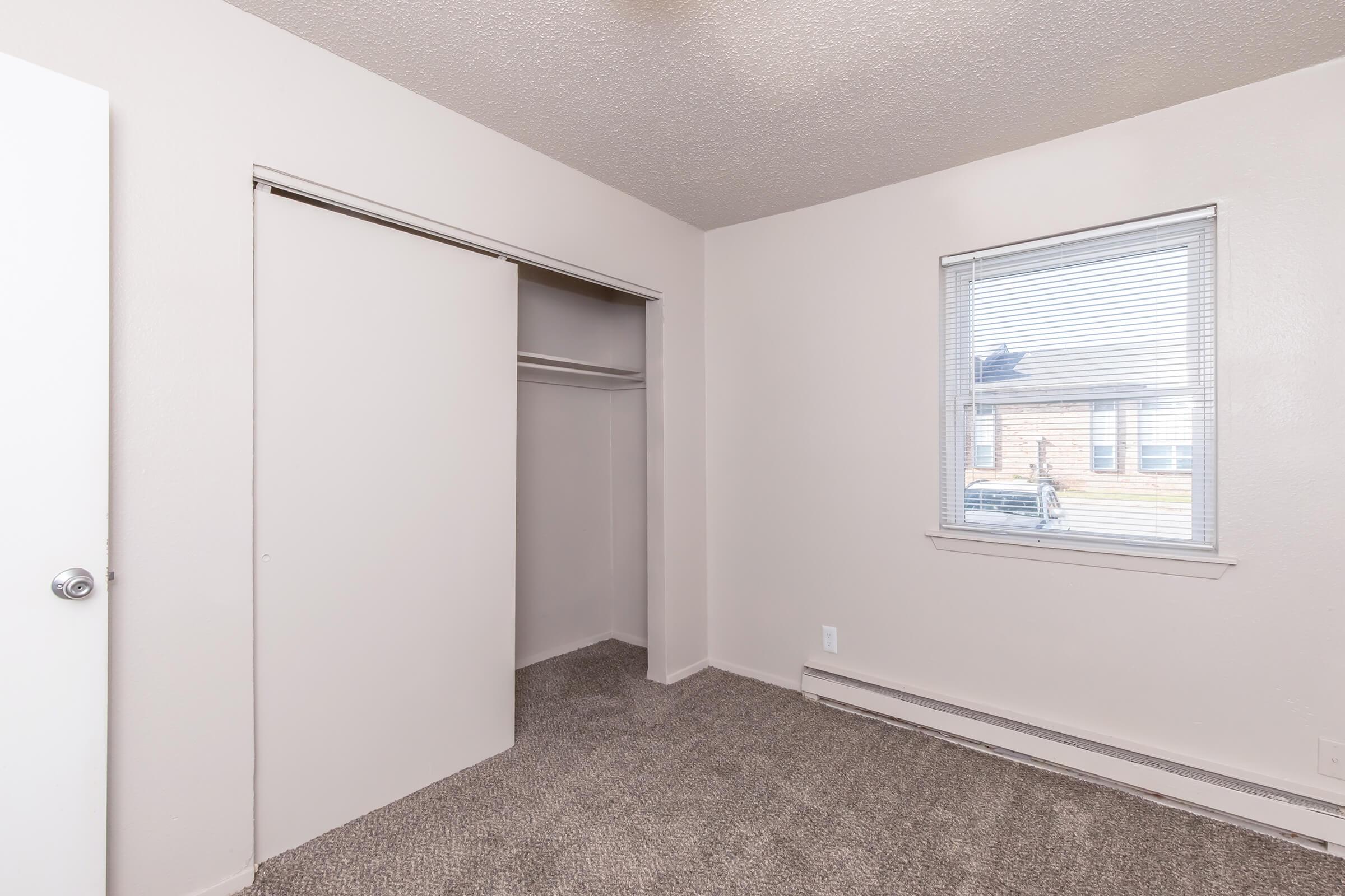 Empty bedroom with light-colored walls and carpeted floor. A closet with sliding doors is visible on the left, and a window with blinds is on the right, allowing natural light in. The room has minimal furnishings, creating a spacious appearance.