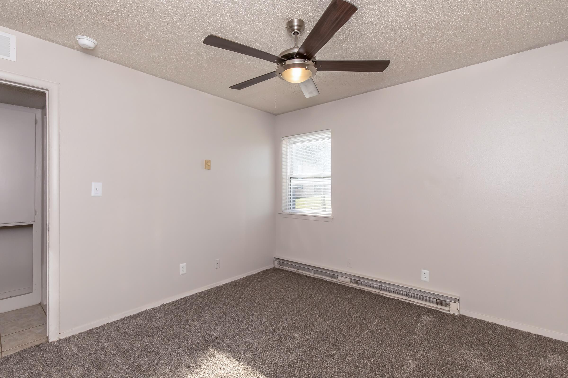 A small, empty room featuring beige walls and a ceiling fan. The floor is covered with a light-colored carpet. A single window allows natural light, and there is a door that likely leads to another room. The overall ambiance is neutral and inviting, suitable for various uses.