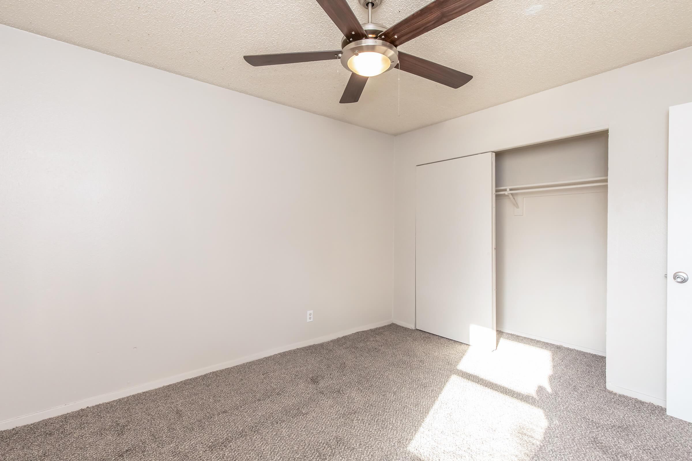 A bright, empty room with light gray walls and a ceiling fan. The floor is covered with light carpet, and there's a closet with sliding doors to the right. Sunlight streams in from a window, creating a warm ambiance. The overall look is clean and minimalistic.