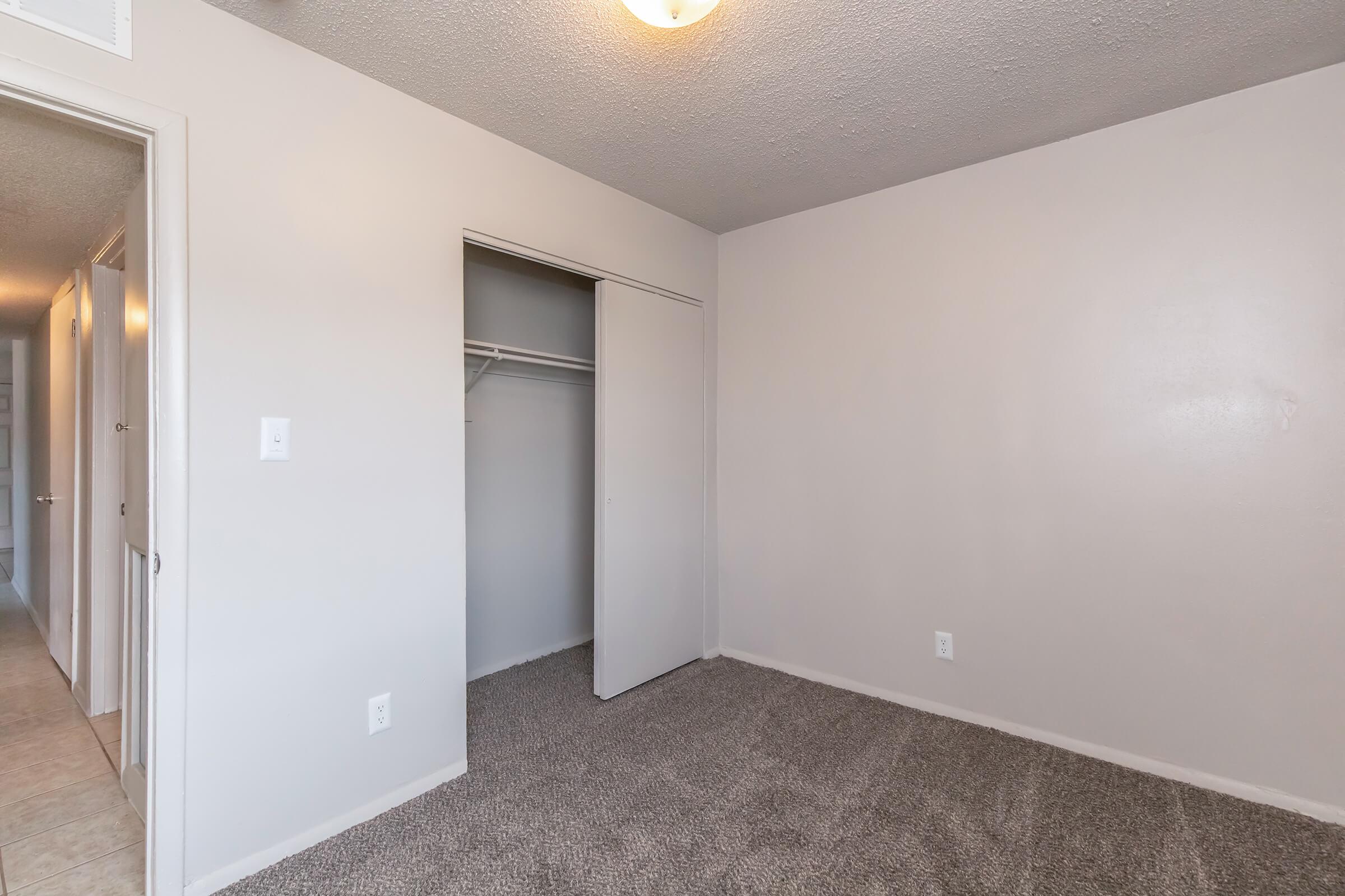 Empty room with light gray walls and carpeted flooring. A closet with sliding doors is visible on the right side. The room has a ceiling light fixture and a doorway leading to another area, suggesting it's part of an apartment or a small living space.