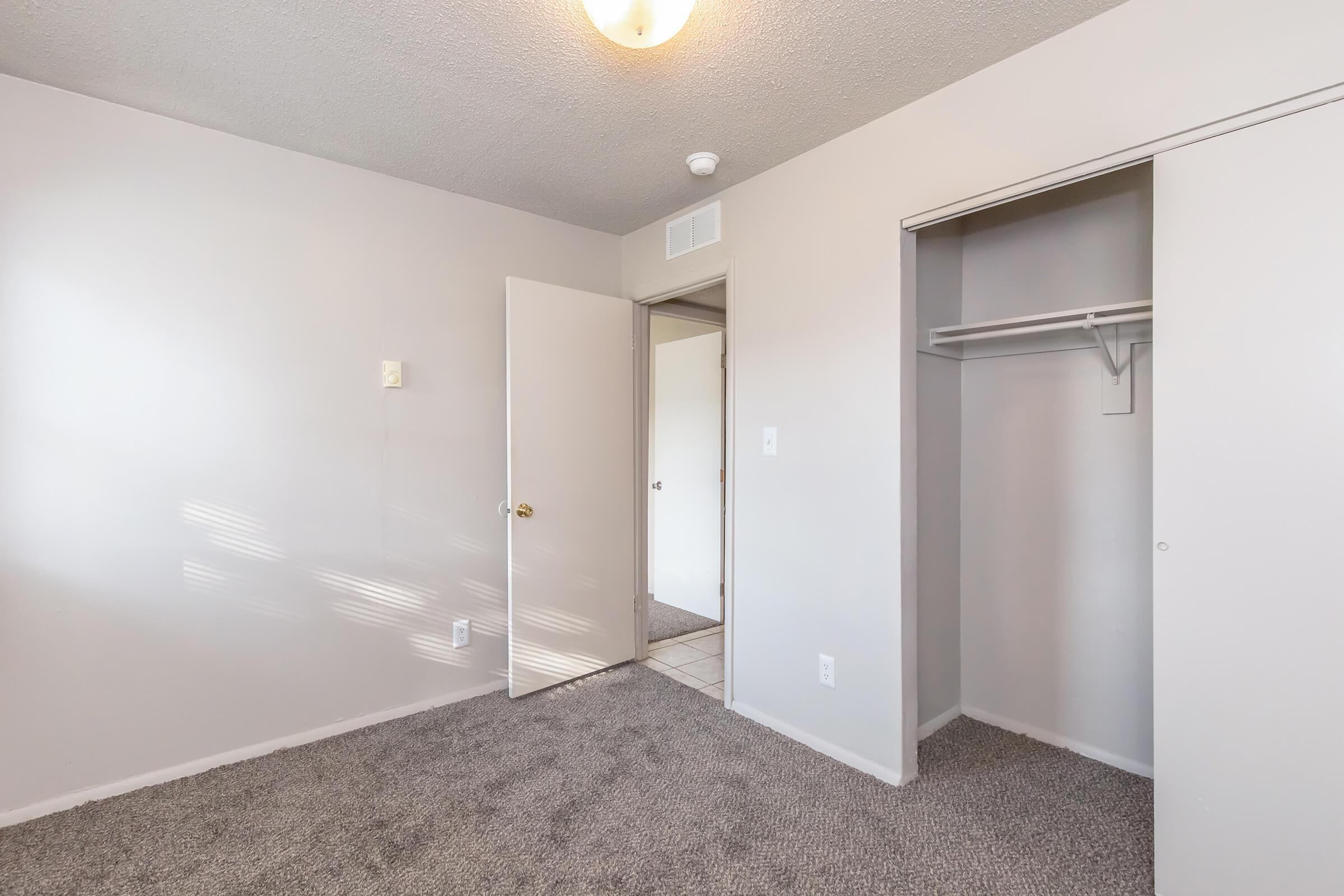 Interior of a small, empty room featuring beige walls, a light fixture on the ceiling, and gray carpet. A door leads to another room, and there is a closet with a single shelf. Natural light casts shadows on the wall, creating a bright, airy atmosphere.
