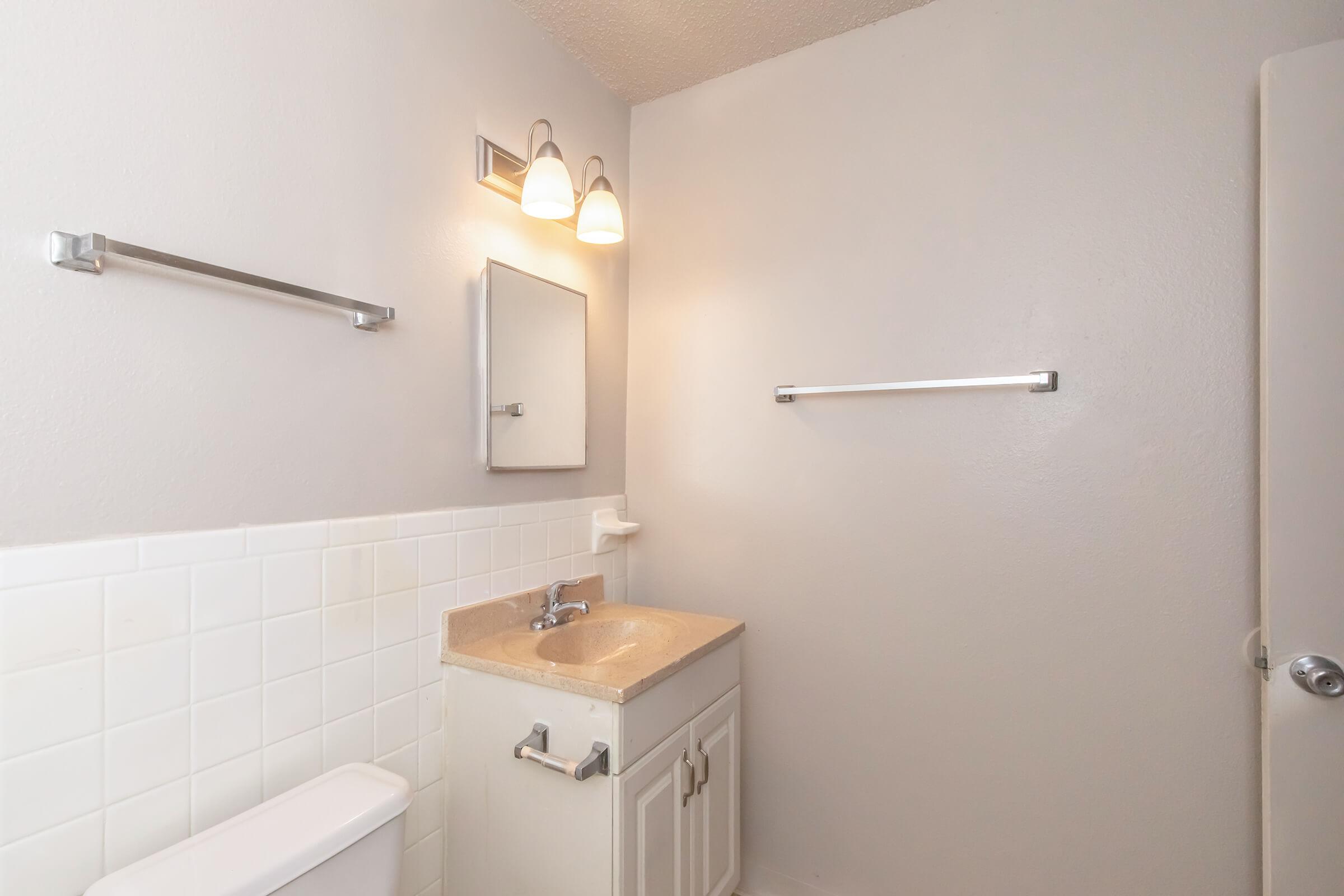 A clean, modern bathroom featuring a white tile floor, a countertop sink with a small mirror above, and two wall-mounted light fixtures. There are towel bars on the walls, and a white toilet is visible. The walls are painted a light gray, creating a bright and airy atmosphere.