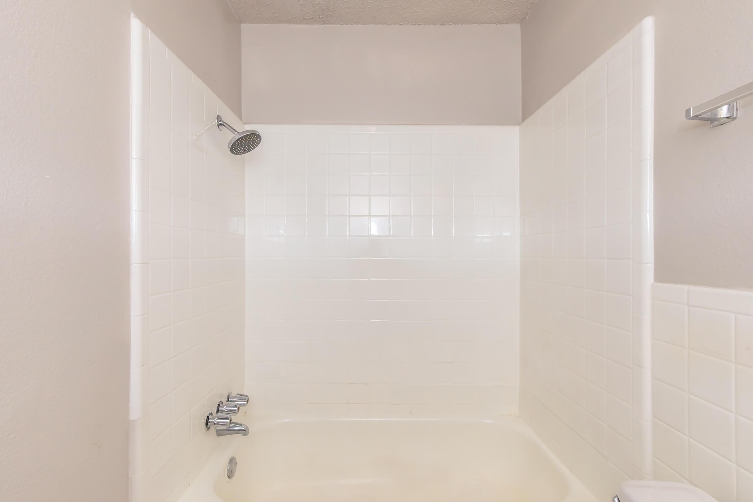A clean and simple bathroom shower space featuring white tiled walls and a beige bathtub. The showerhead is mounted above, and there are controls for hot and cold water. The overall design is minimalistic and uncluttered, with neutral colors and natural light enhancing the space.