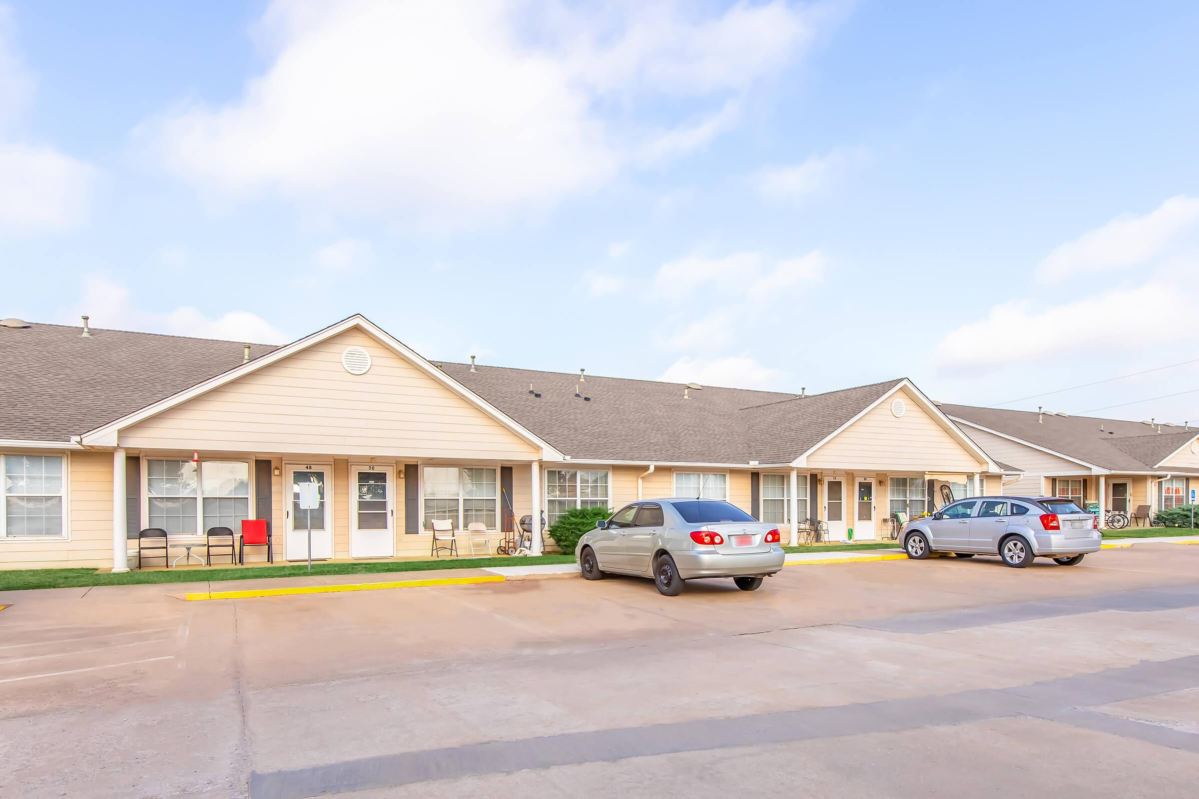 A row of single-story residential apartments with beige siding, featuring front porches and windows. two parked cars are visible in the foreground, along with green grass and a clear blue sky. Ideal for showcasing multi-family housing options.