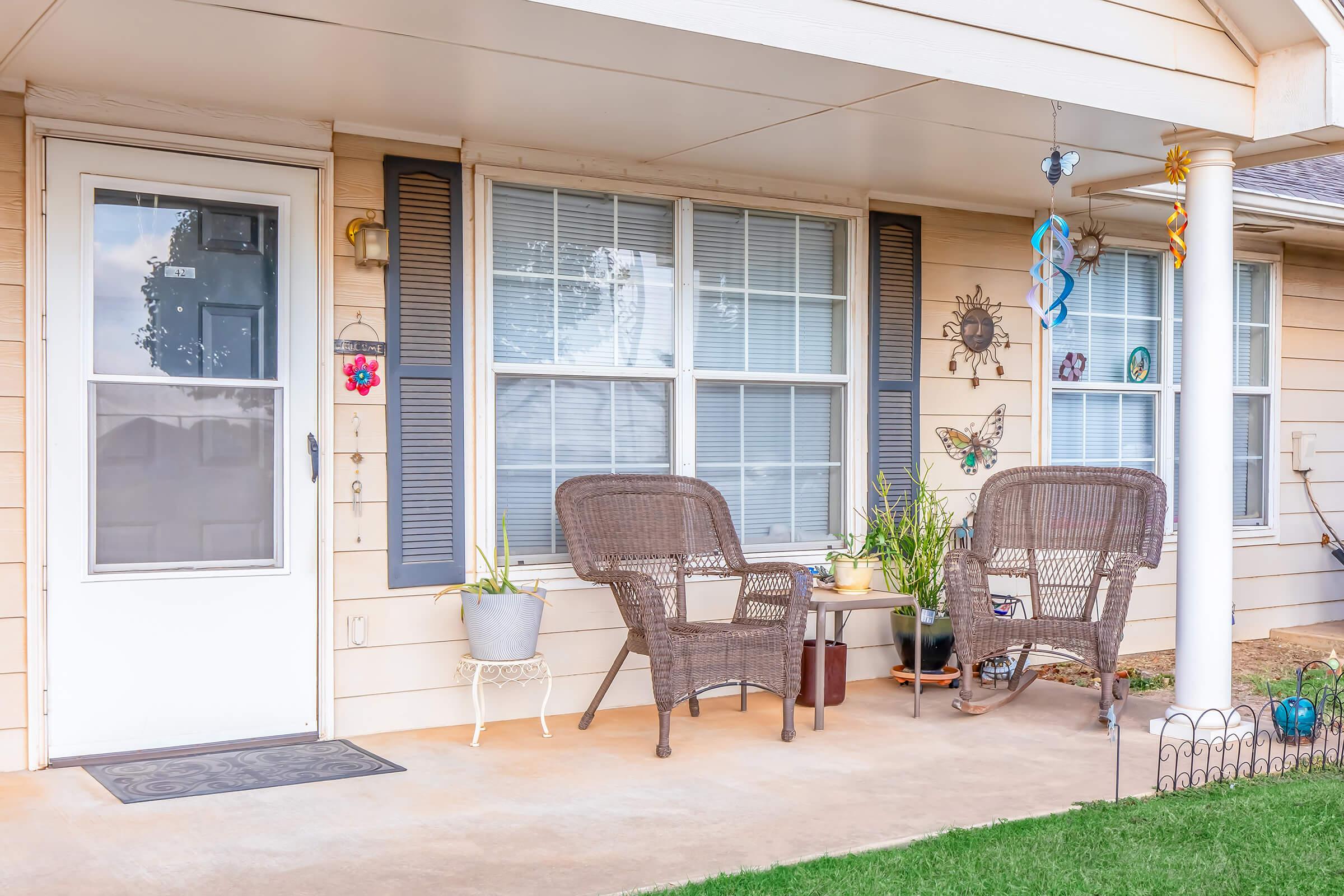 A cozy front porch featuring two wicker chairs, a small table, and potted plants. The door has a screen and decorative elements such as wall hangings and windchimes. The ground is concrete and there is a small patch of grass in front of the porch, creating a welcoming outdoor space.