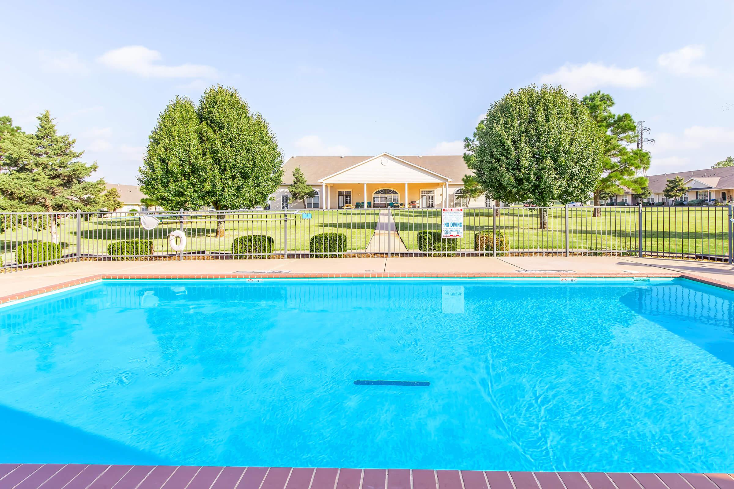 A clear, inviting swimming pool in the foreground with a well-maintained grassy area and trees surrounding it. In the background, a building with a patio and large windows is visible, set against a blue sky. The pool area is enclosed by a fence, enhancing the serene atmosphere.