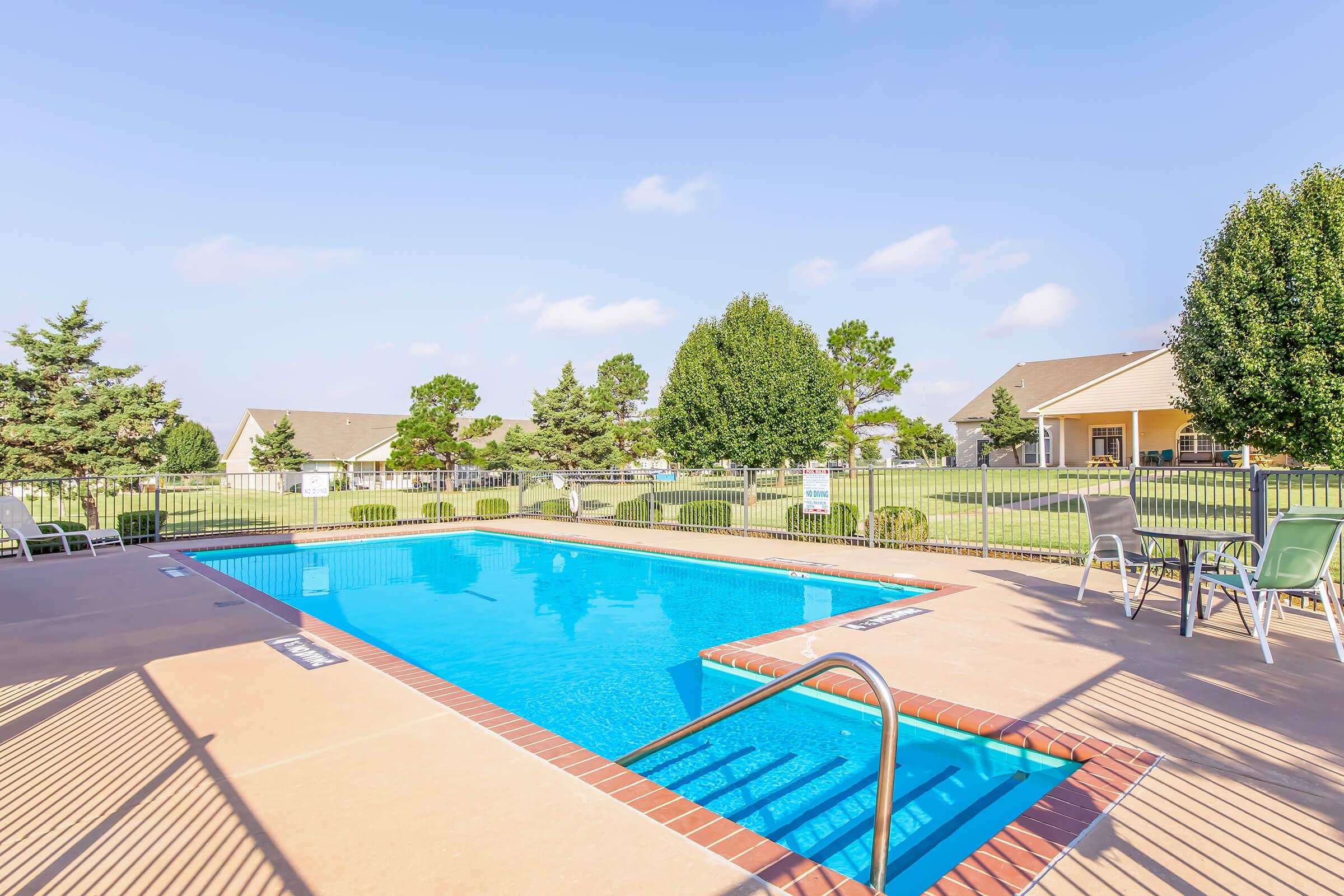 A clear blue swimming pool surrounded by a tan patio. Green trees and well-maintained lawns are visible in the background, with a few houses in view. White lounge chairs line the poolside, and a set of steps leads into the water, inviting for a refreshing swim.
