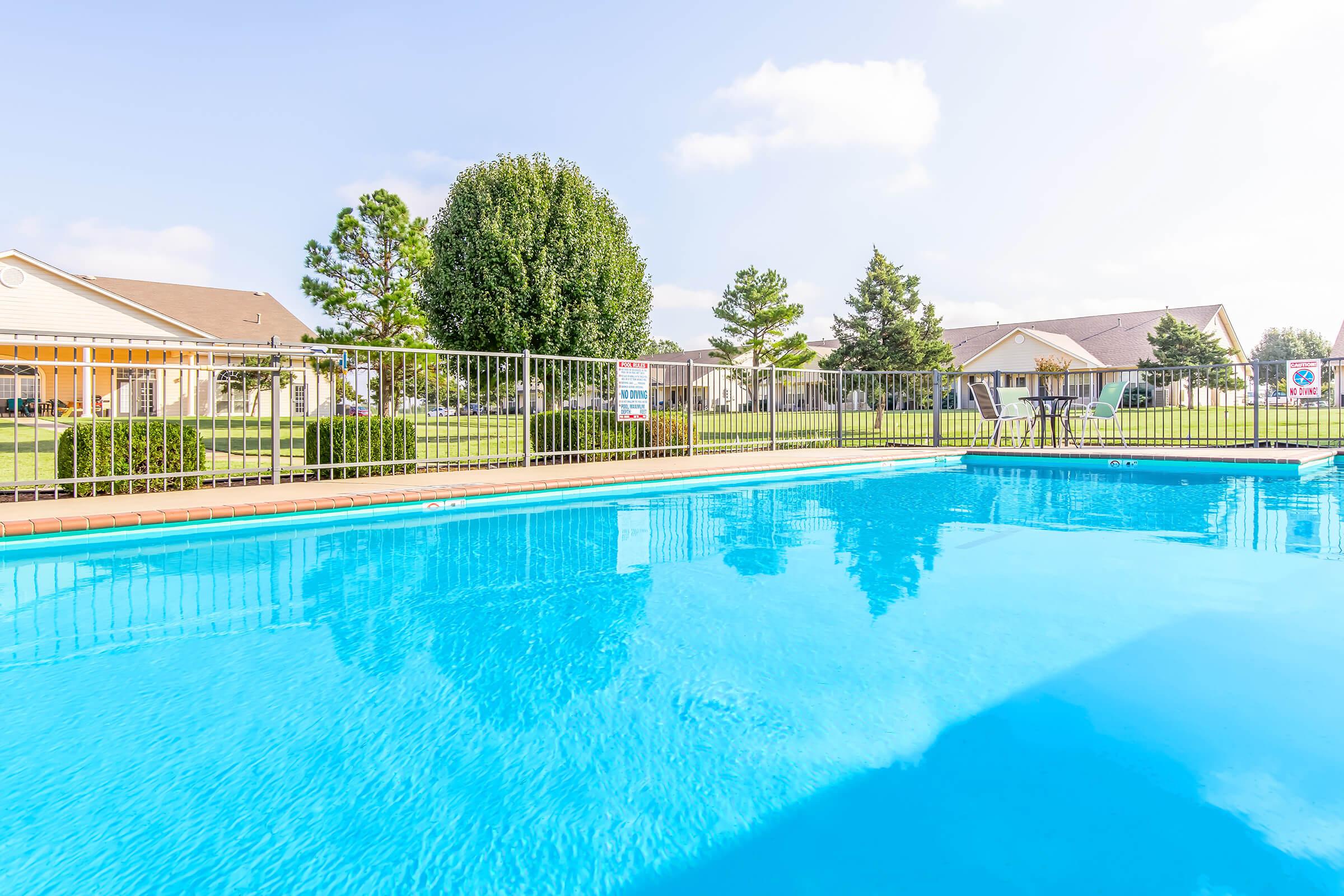 A clear, inviting swimming pool surrounded by a metal fence, with lush green grass and trees in the background. Residential buildings are visible in the distance under a sunny sky, creating a relaxing outdoor atmosphere. The water reflects the blue sky, enhancing the serene environment.