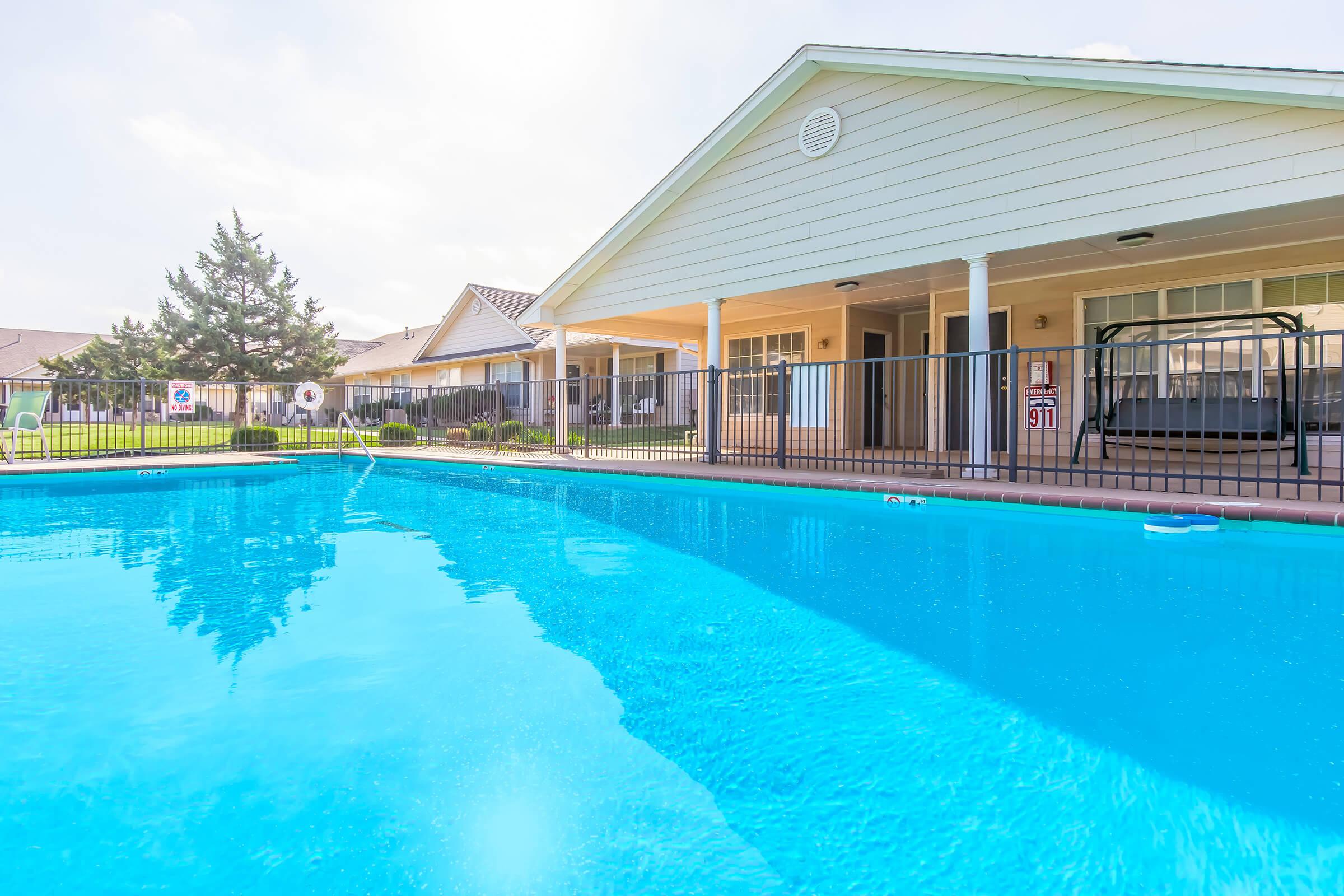 A clear blue swimming pool in the foreground, with the sun reflecting on the water. In the background, there are residential buildings with porches and a well-maintained lawn. A fence surrounds the pool area, adding privacy to the space, and the sky is bright and partially cloudy.