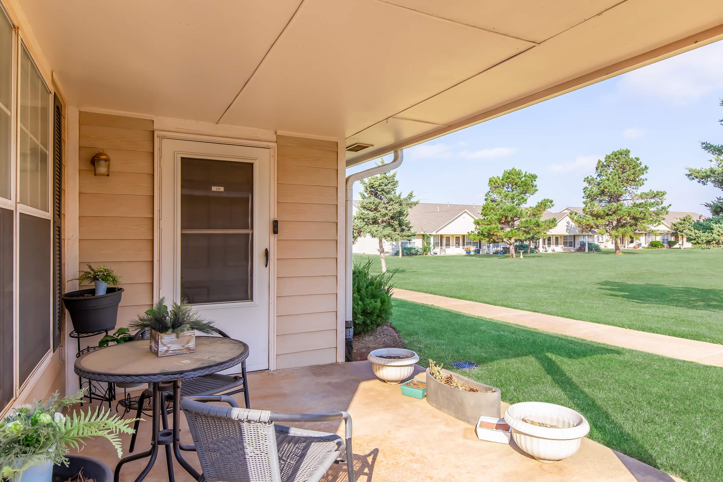 A cozy porch with a small round table and chairs, surrounded by potted plants. A door leads into the house, and a spacious green lawn stretches out in the background, dotted with trees and houses. The scene is bright and inviting, suggesting a peaceful outdoor space.