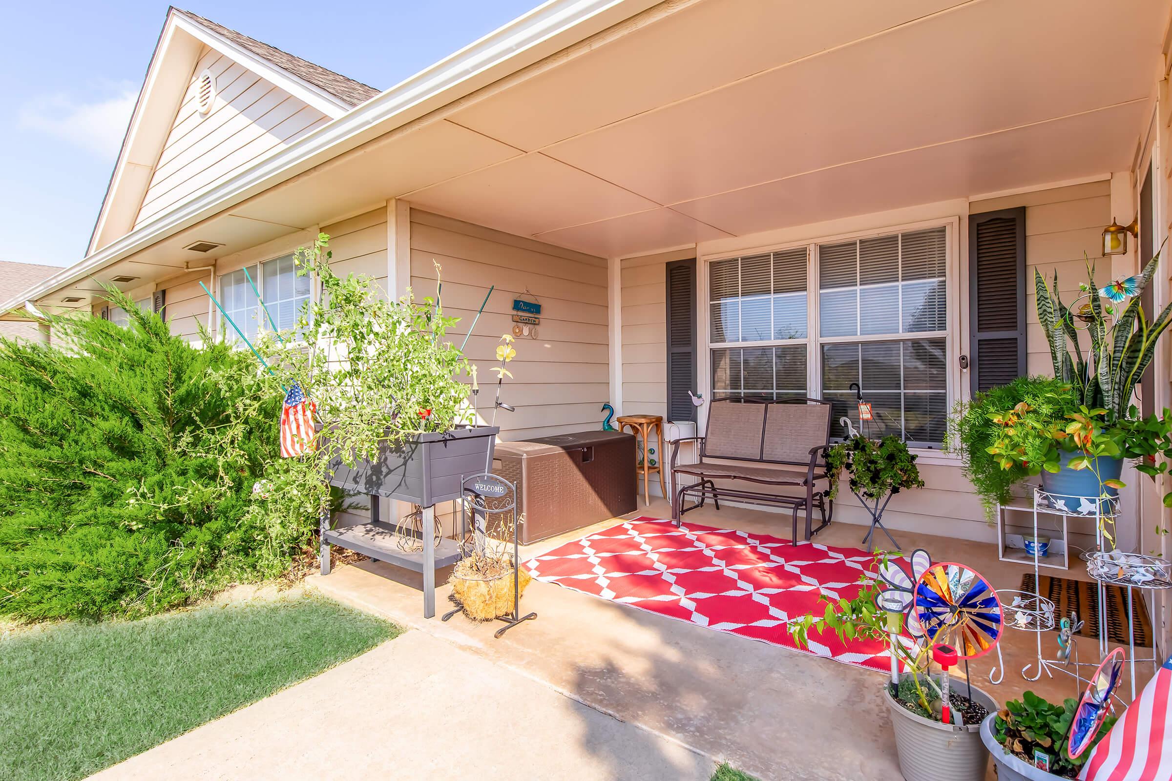 A welcoming front porch featuring a patterned red rug, a comfortable bench, potted plants, and decorative items like a pinwheel. A planter and vibrant greenery enhance the cozy atmosphere, with a clear sky visible in the background. Ideal for relaxation and enjoying the outdoors.