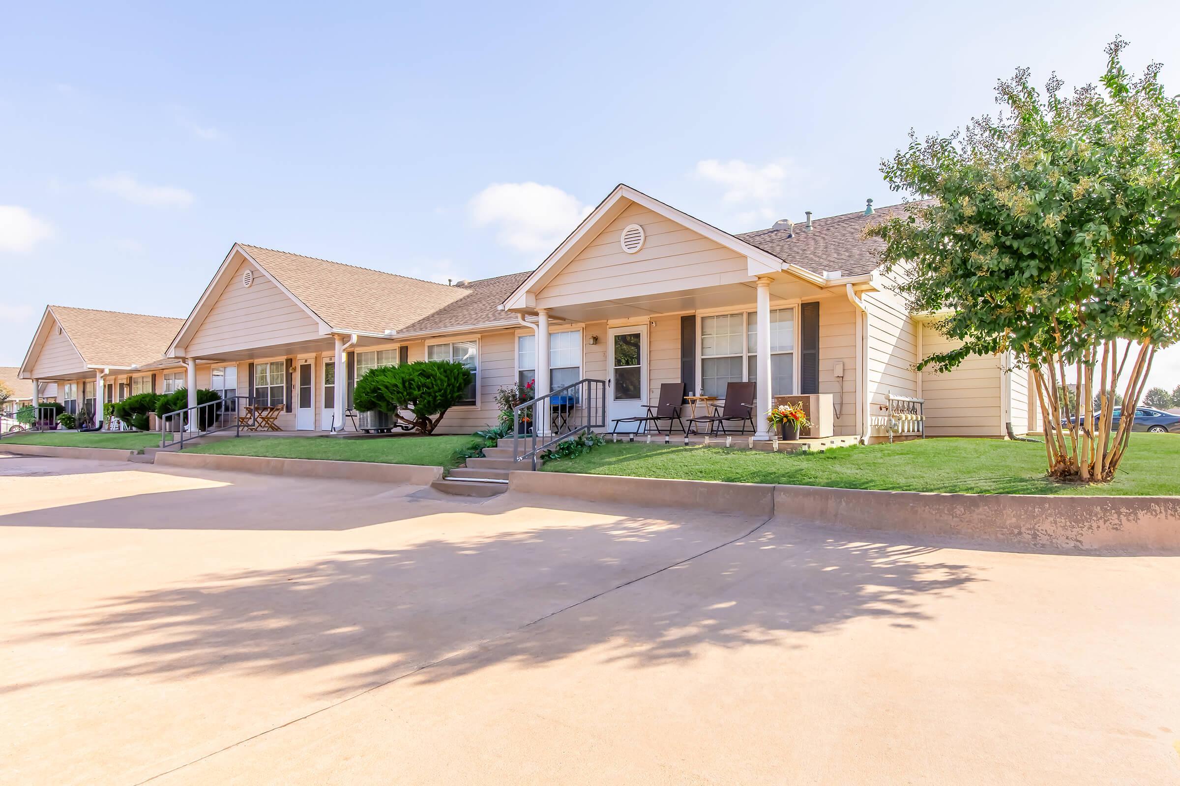 Rows of single-story beige cottages with sloped roofs, each featuring a porch and surrounding greenery. The pathway is paved, leading to the front steps of the homes. Bright blue sky above with a few fluffy clouds enhances the inviting atmosphere.