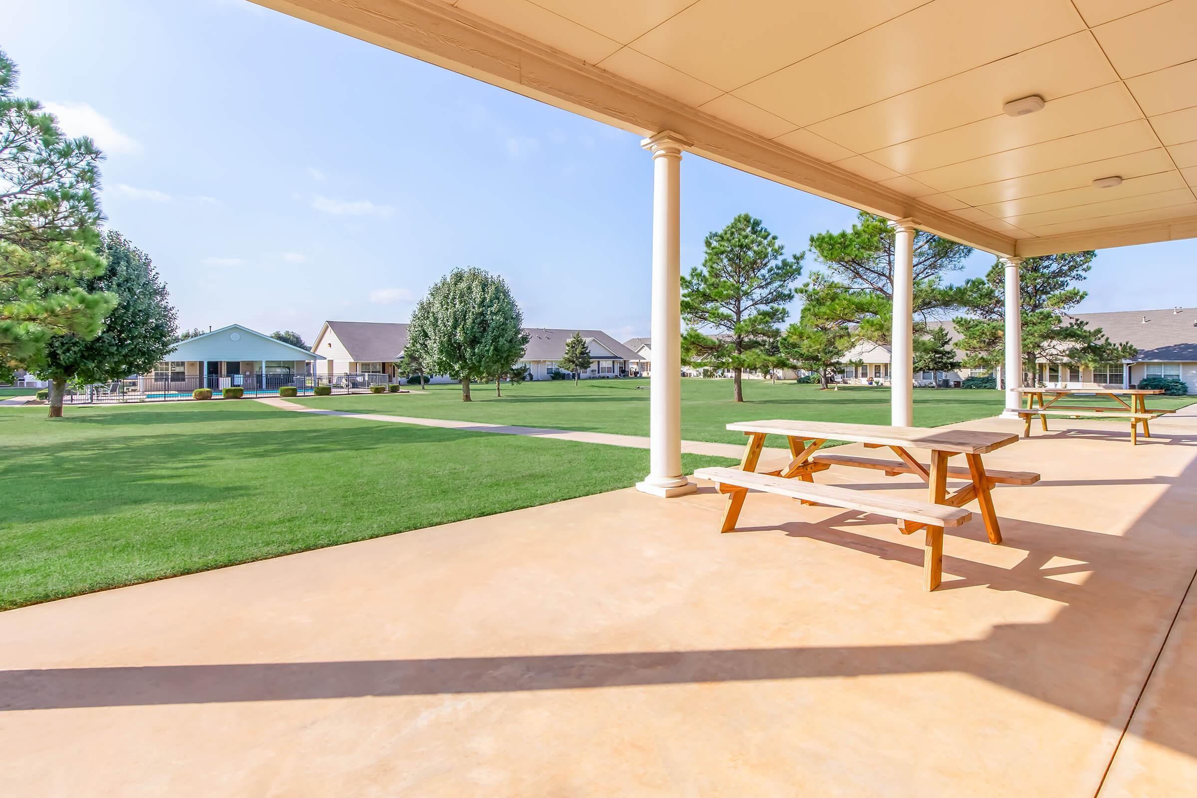 View from a covered patio showcasing a green lawn with trees and several residential buildings in the background. Two picnic tables are positioned on the patio, providing a space for outdoor relaxation. The scene is bright and sunny, highlighting a peaceful community atmosphere.