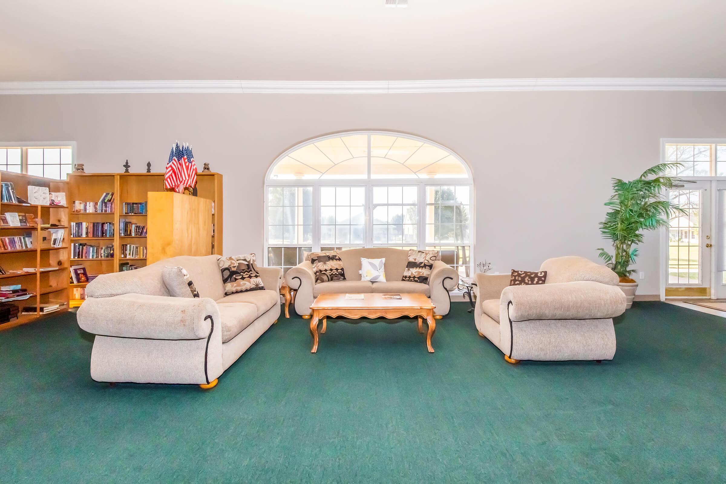 A cozy living room featuring two light-colored sofas with decorative pillows, a wooden coffee table in the center, and a large window in the background allowing natural light. There's a bookshelf filled with books on one side and a potted plant, creating a warm and inviting atmosphere.