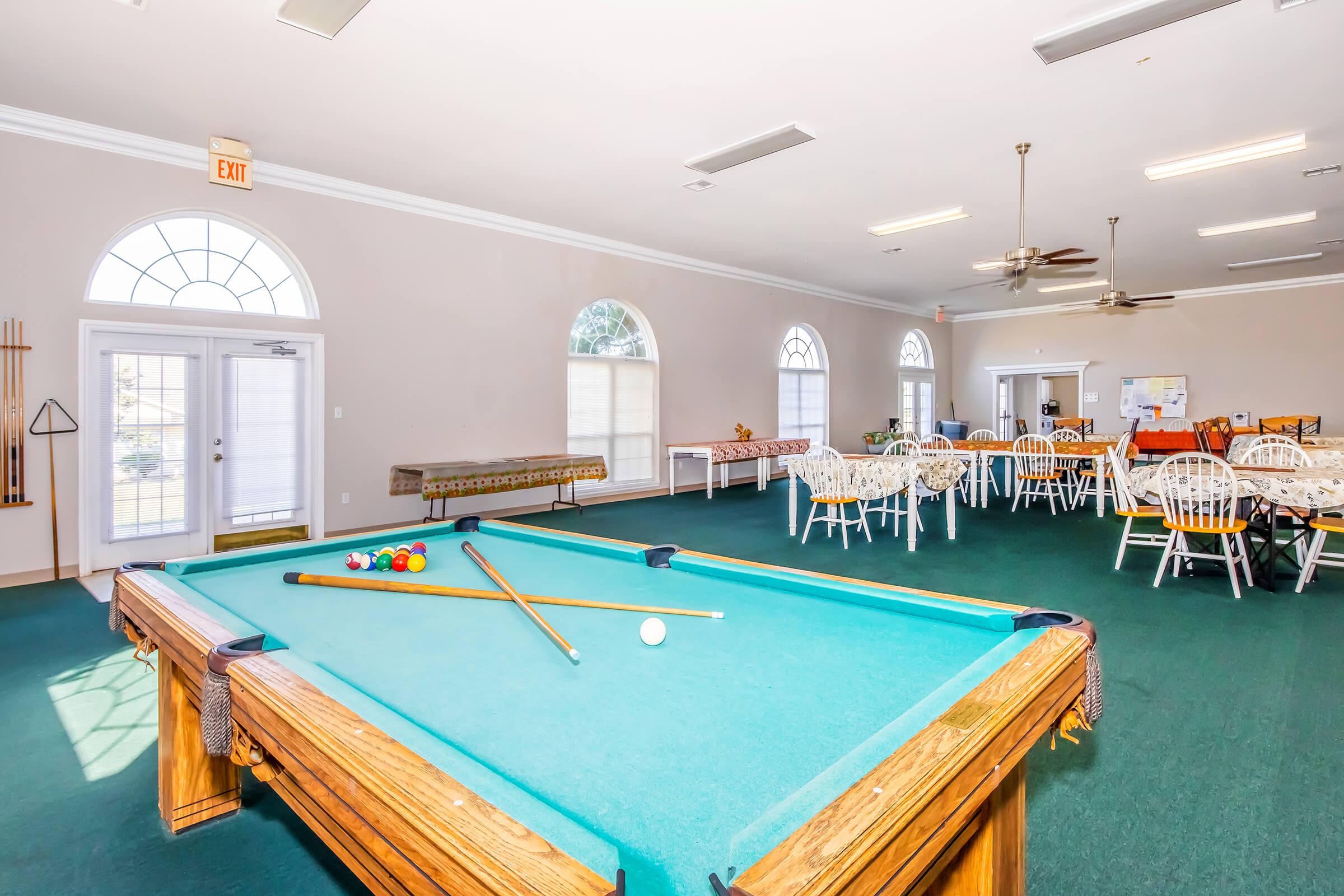 A spacious game room featuring a green felt pool table with two cues and colored balls, surrounded by a dining area with white wooden chairs and round tables. Large windows provide natural light, and the room has a clean, inviting atmosphere, suitable for recreation and social gatherings.