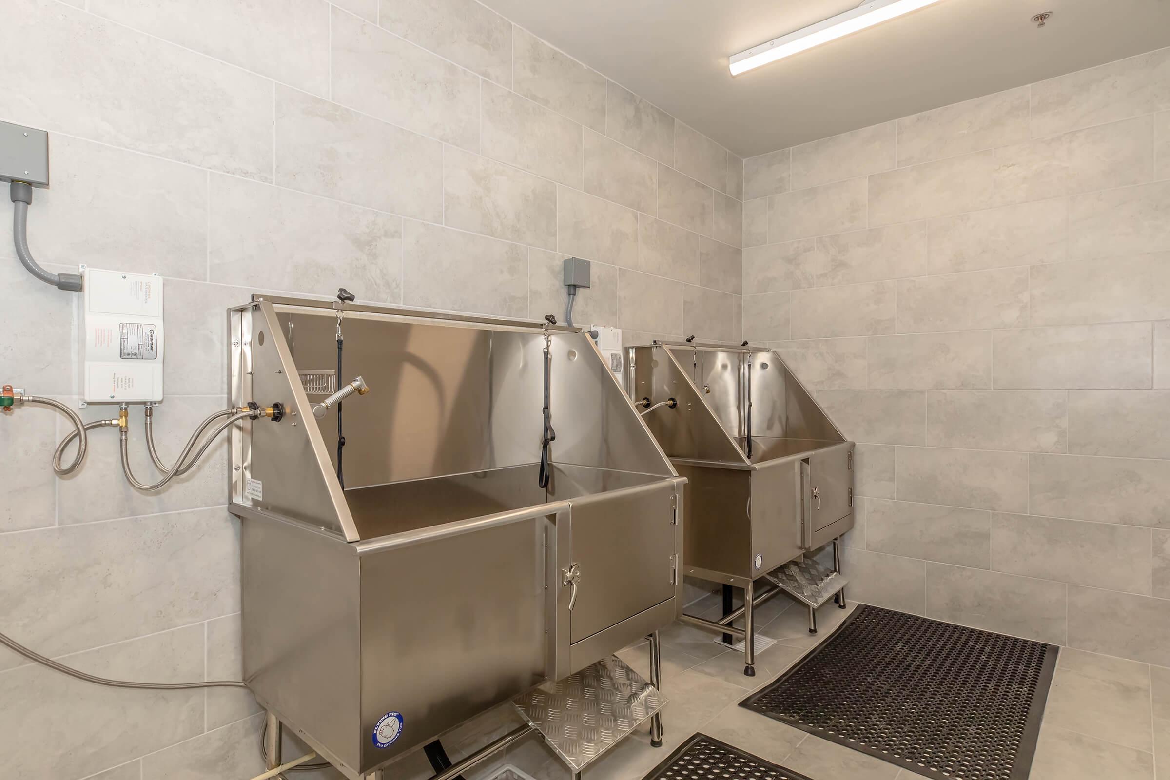 Two stainless steel pet bathing stations in a tiled room, equipped with hoses and drainage. The area features a clean, modern design with gray tiles and rubber mats for safety. Bright overhead lighting provides a well-lit space for grooming.