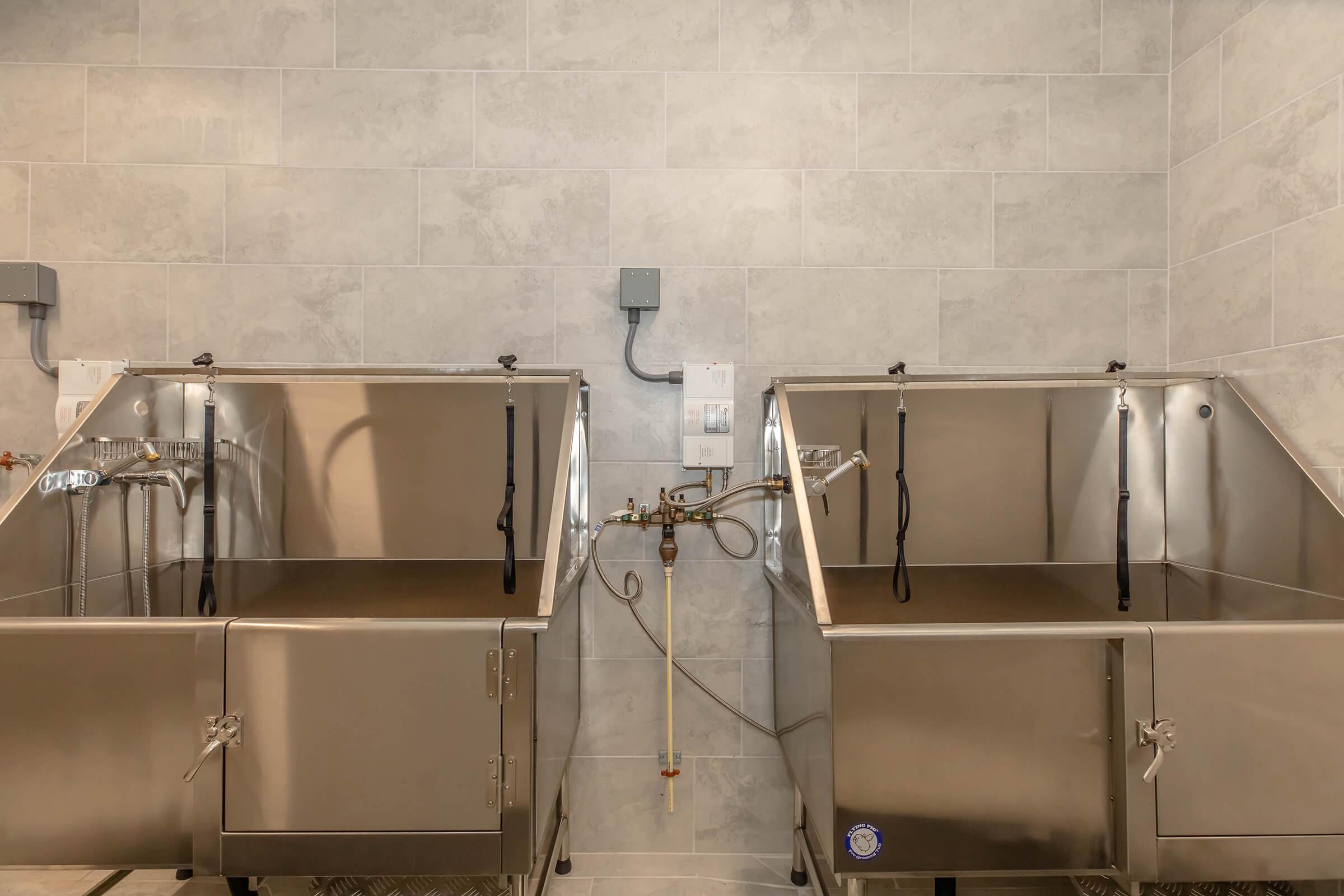 Two stainless steel wash basins with faucets, set against a tiled wall. Each basin has a unique design, with one featuring a hose attachment and a control panel on the wall. The interior is empty, and the overall aesthetic is clean and modern.