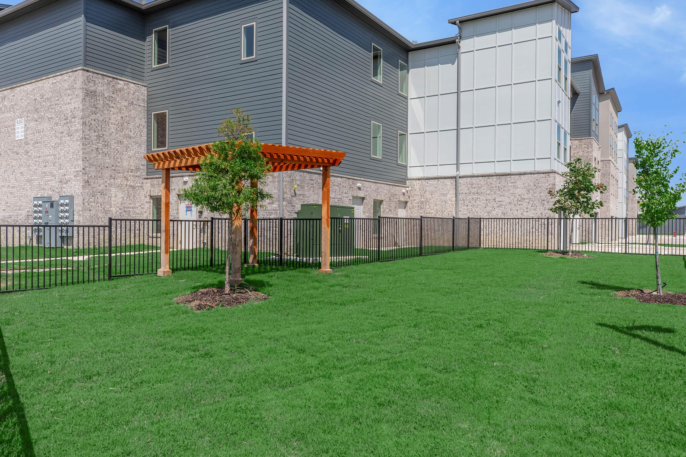 A grassy area with a small pergola and trees, fenced in, adjacent to a modern multi-story building. The building features a combination of brick and gray siding. Utility boxes are visible near the fence, and the sky is clear and blue.