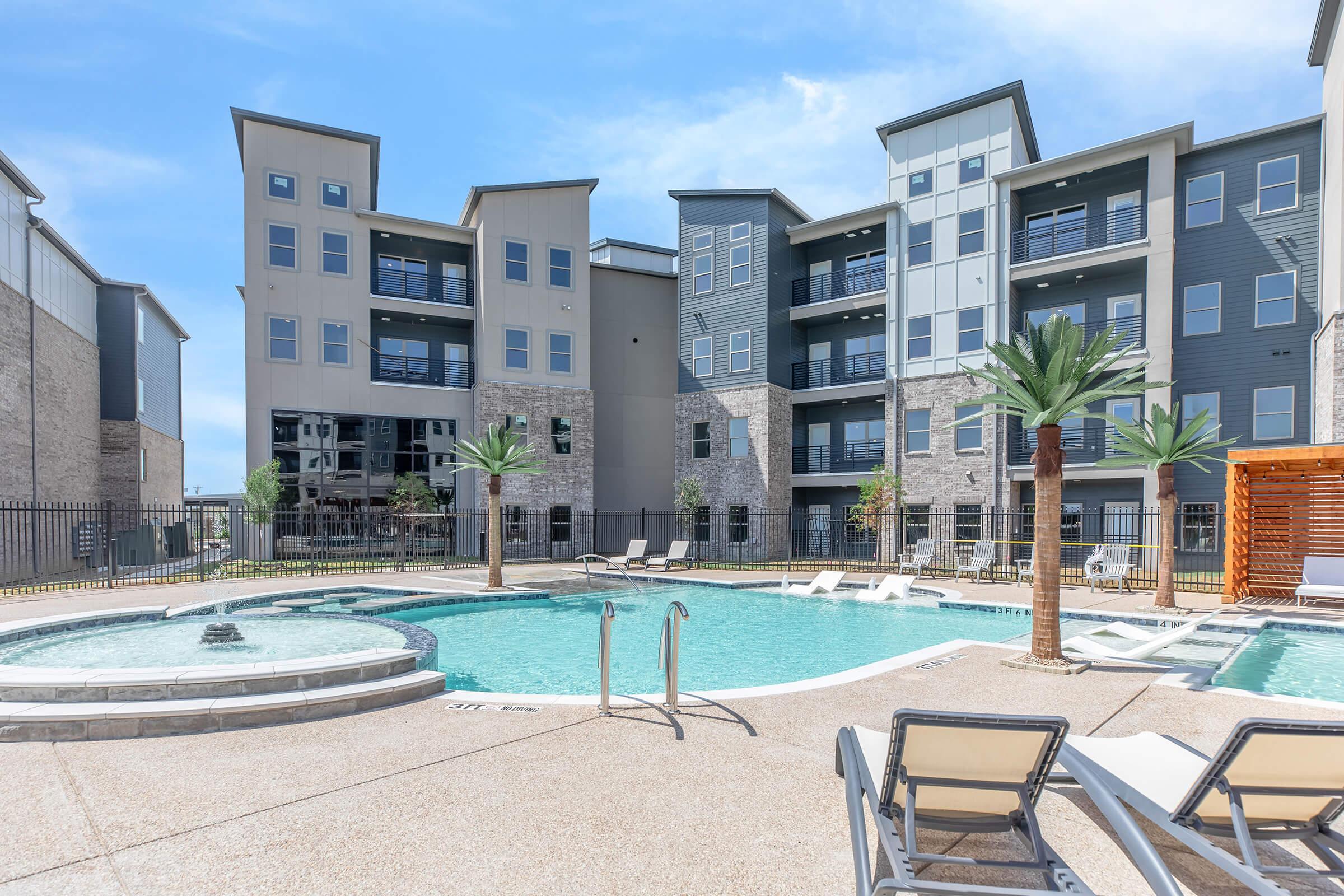 A sunny outdoor pool area surrounded by residential buildings. The pool features a fountain and is lined with lounge chairs and palm trees. Modern apartments with large windows provide a contemporary backdrop, and a shaded seating area is visible to the side. Clear blue skies enhance the inviting atmosphere.