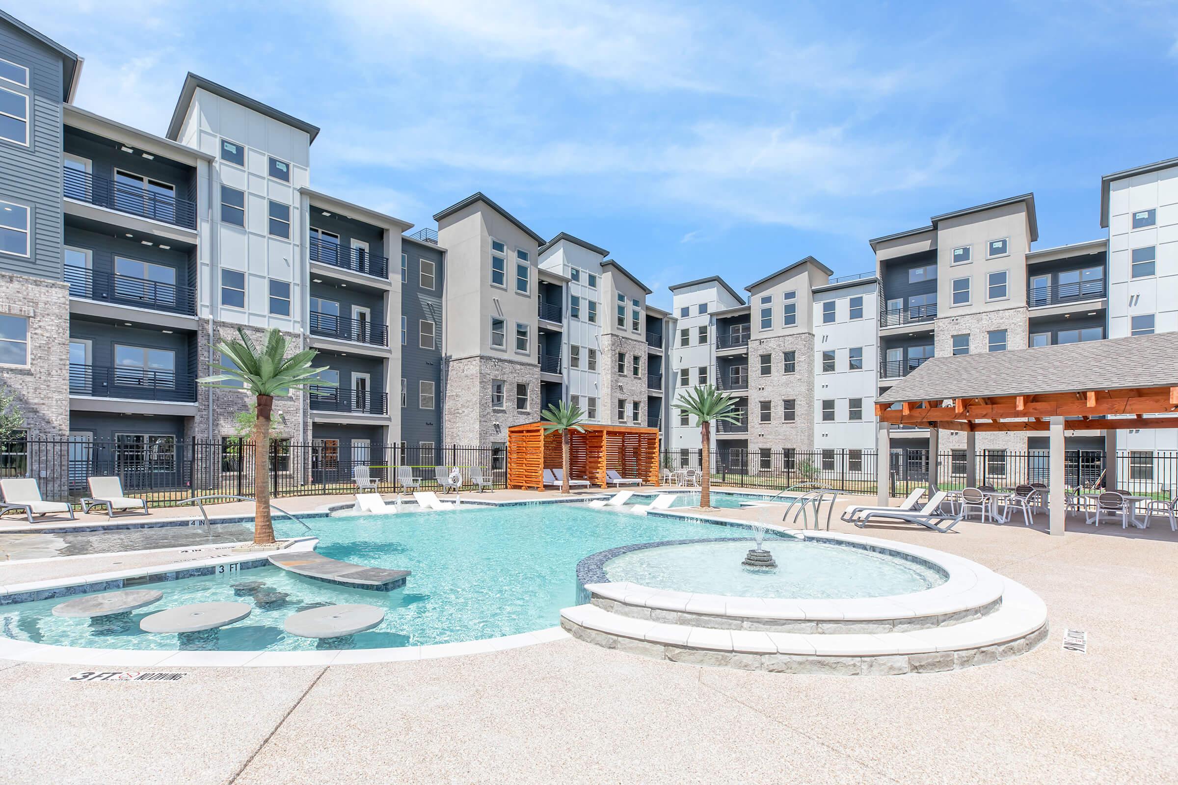 A large outdoor swimming pool surrounded by lounge chairs and palm trees, with modern apartment buildings in the background. There is a wooden pergola nearby, and clear blue skies above, creating a relaxing atmosphere.