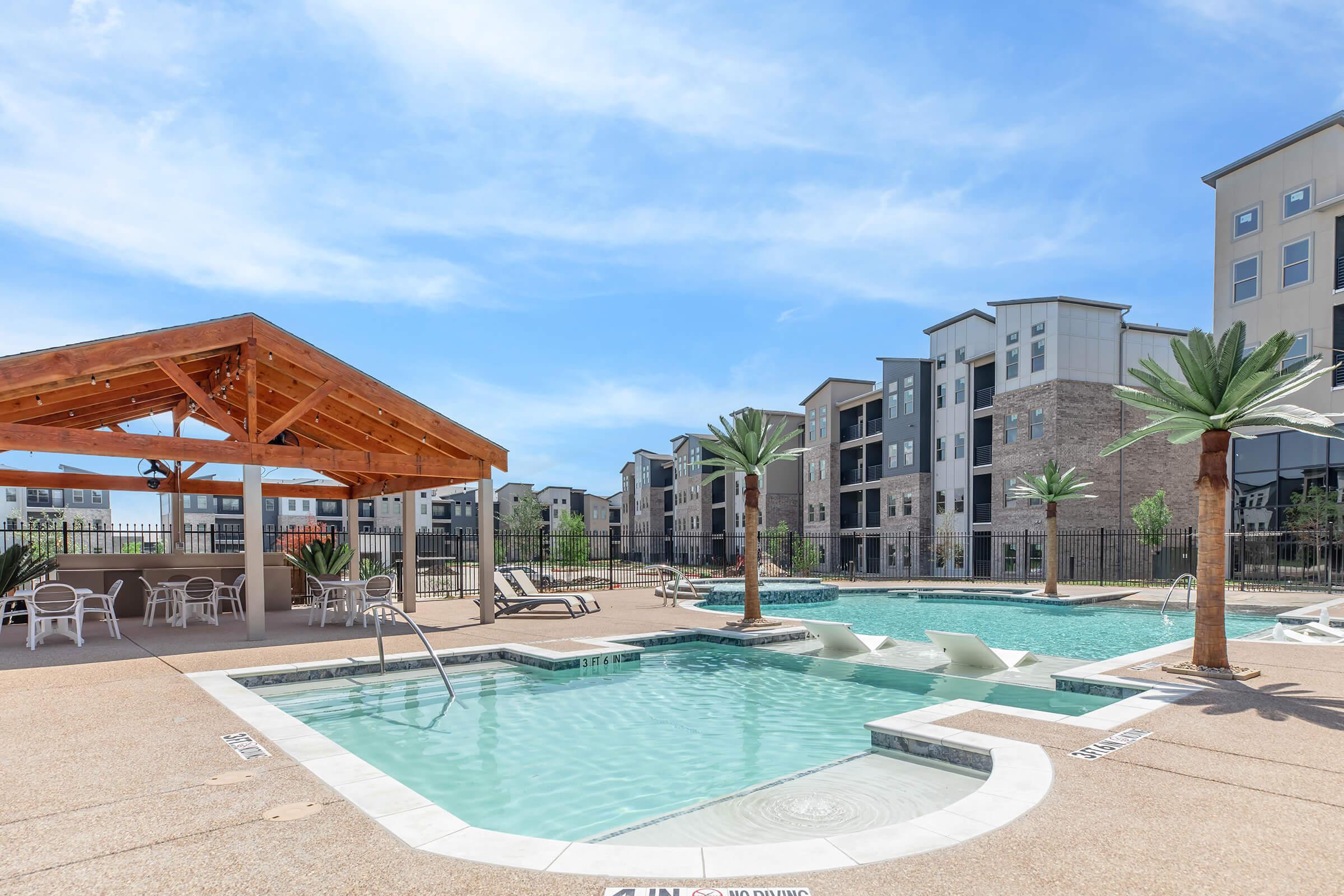 A view of a swimming pool area featuring a hot tub, lounge chairs, and palm trees. In the background, modern apartment buildings are visible under a clear blue sky. A shaded gazebo with seating is also present beside the pool. The setting is bright and inviting, suggesting a relaxing outdoor space.