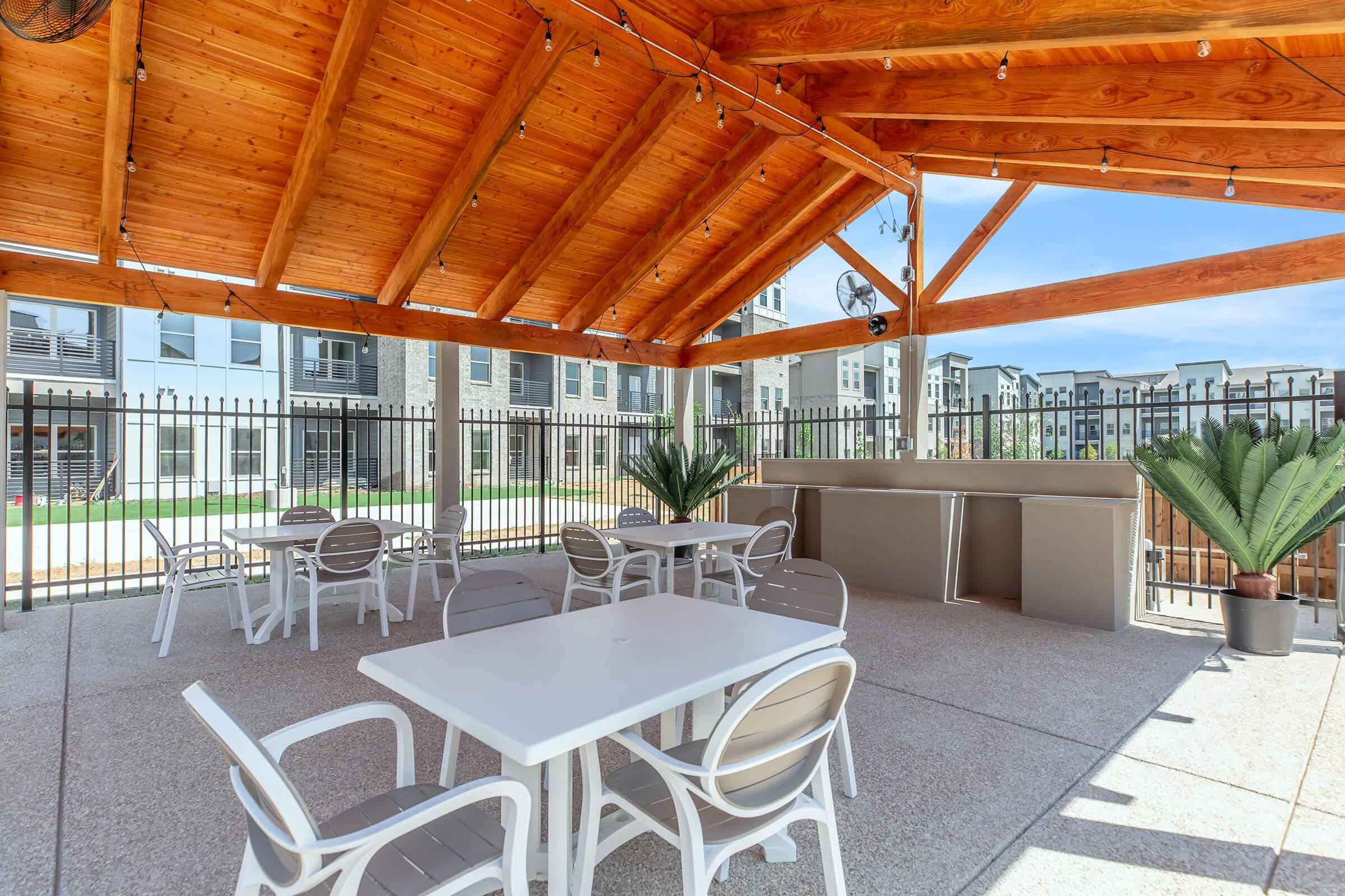 A shaded outdoor seating area with white tables and chairs underneath a wooden pergola. The space features potted plants and is enclosed by a black fence. In the background, there are modern apartment buildings and green lawn areas, creating a welcoming environment for relaxation or social gatherings.