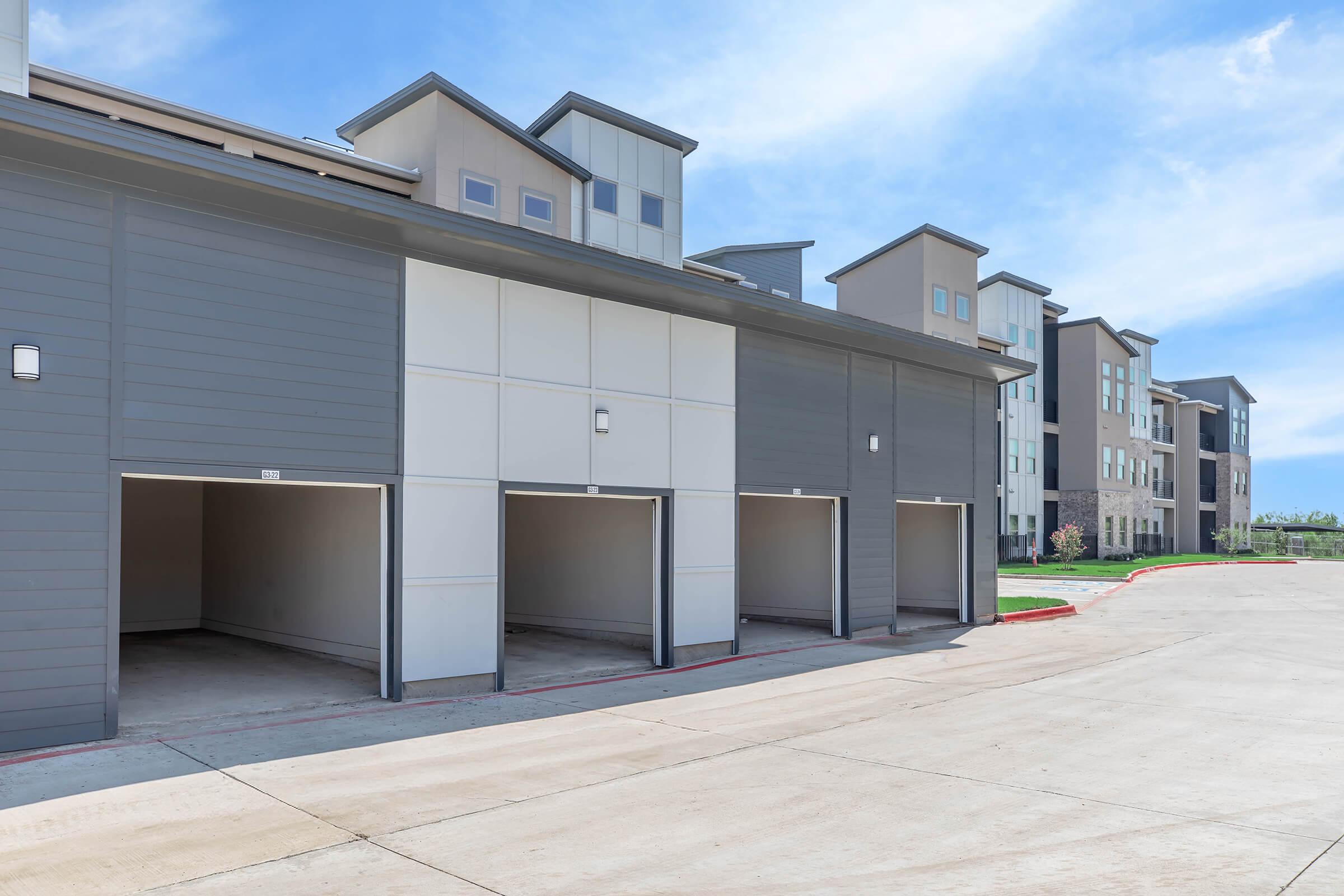 A row of empty garage doors attached to a modern multi-story apartment building. The garages are lined up against a paved area with a clear blue sky overhead, showcasing a contemporary architectural design with a mix of gray and beige colors.