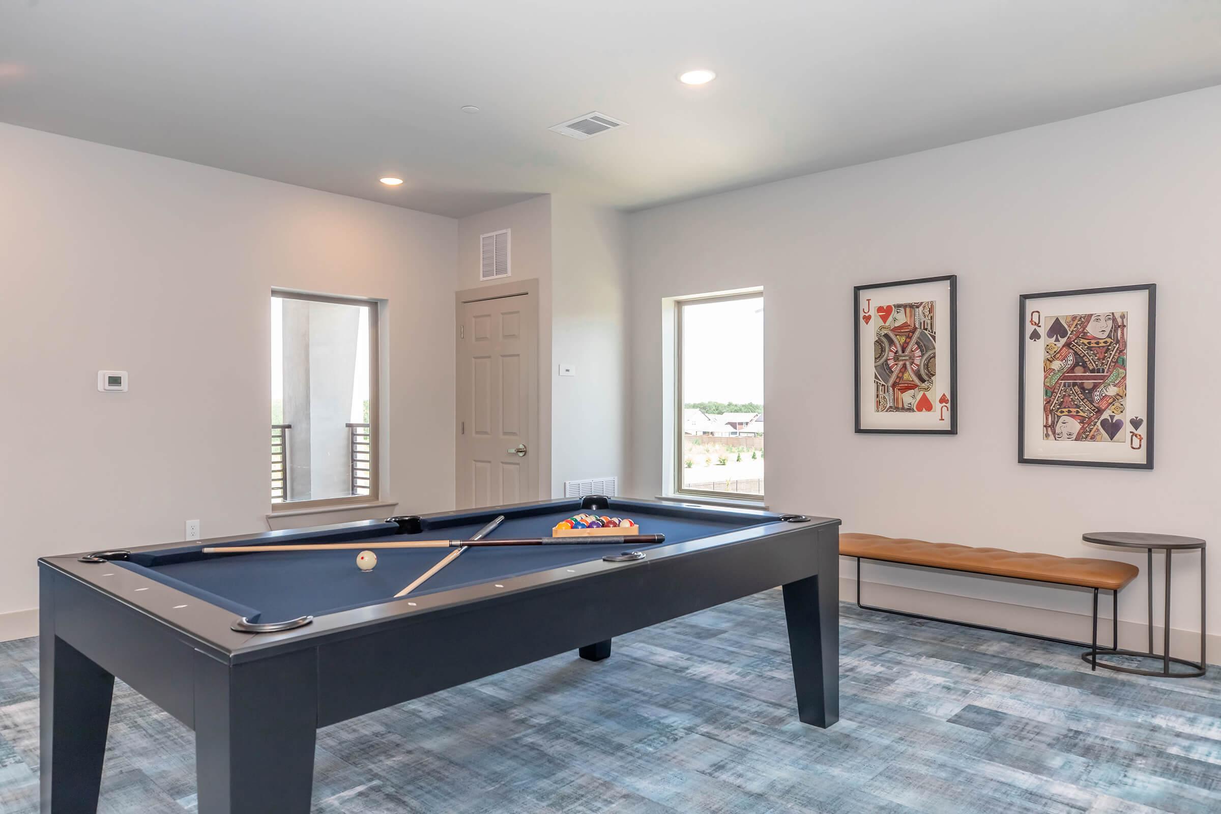 A spacious game room featuring a blue pool table with balls arranged on it, a wooden bench against the wall, and two framed artworks. Large windows allow natural light to fill the room, with modern decor and carpet enhancing the inviting atmosphere.