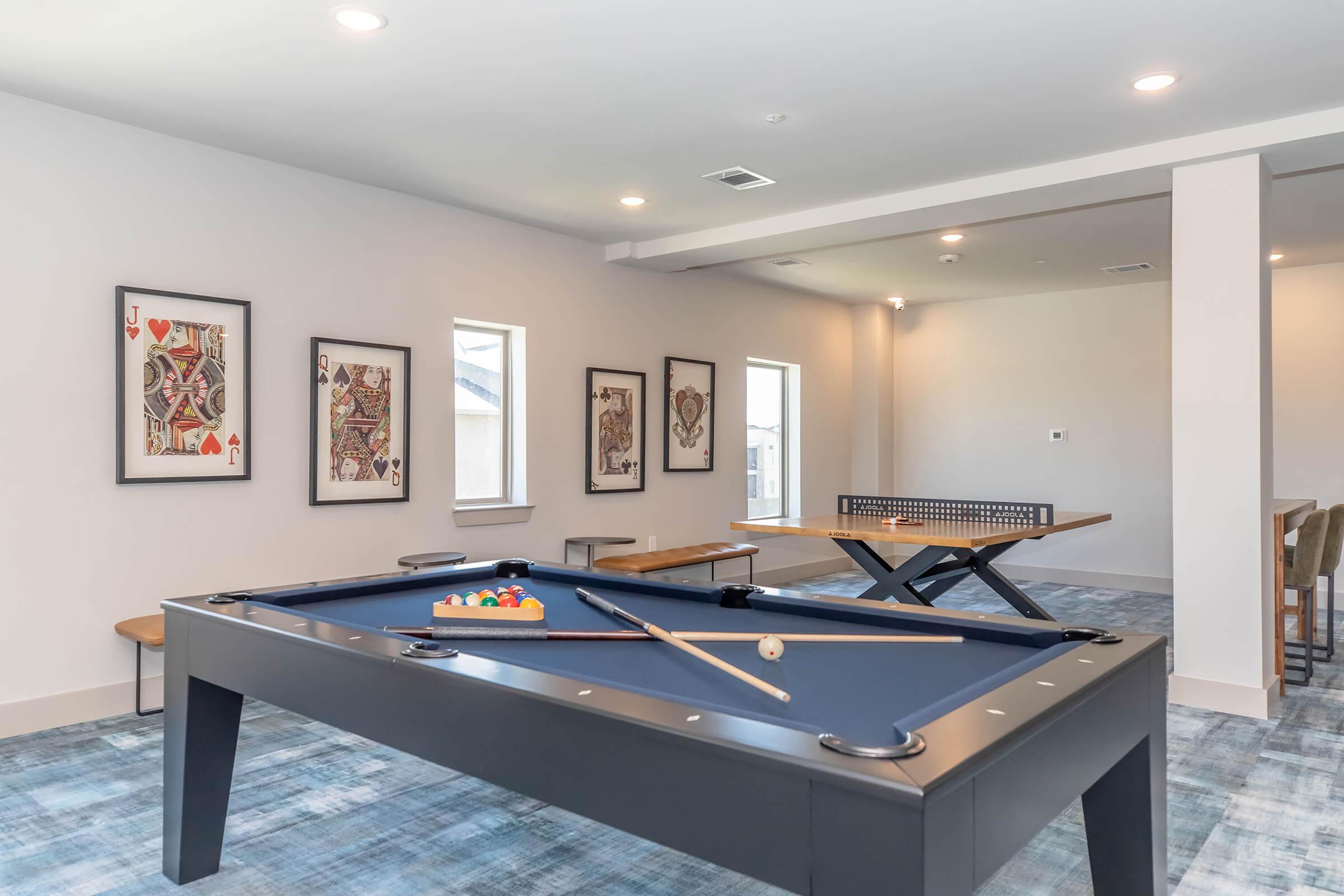 A spacious game room featuring a blue pool table with cues and a set of balls, alongside a ping pong table and modern artwork displayed on the walls. The room is brightly lit with natural light coming through windows and has a contemporary design.