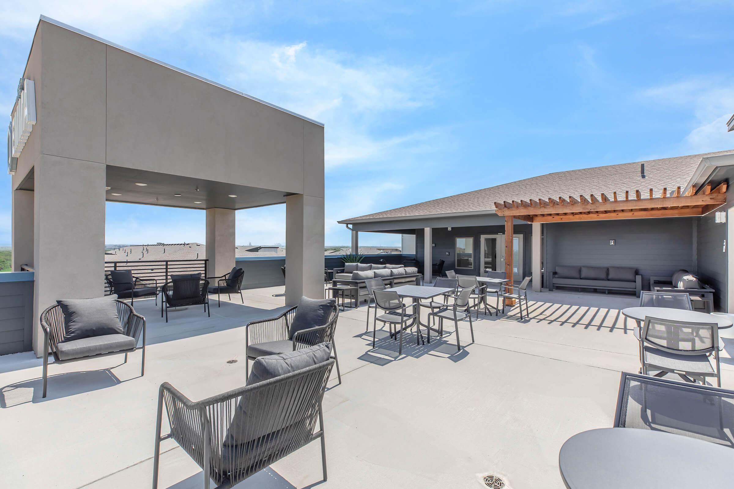 Rooftop patio with modern outdoor furniture, including tables and chairs. There is a shaded area with a pergola, and a clear blue sky is visible. The space is designed for relaxation and social gatherings, showcasing a contemporary architectural style.