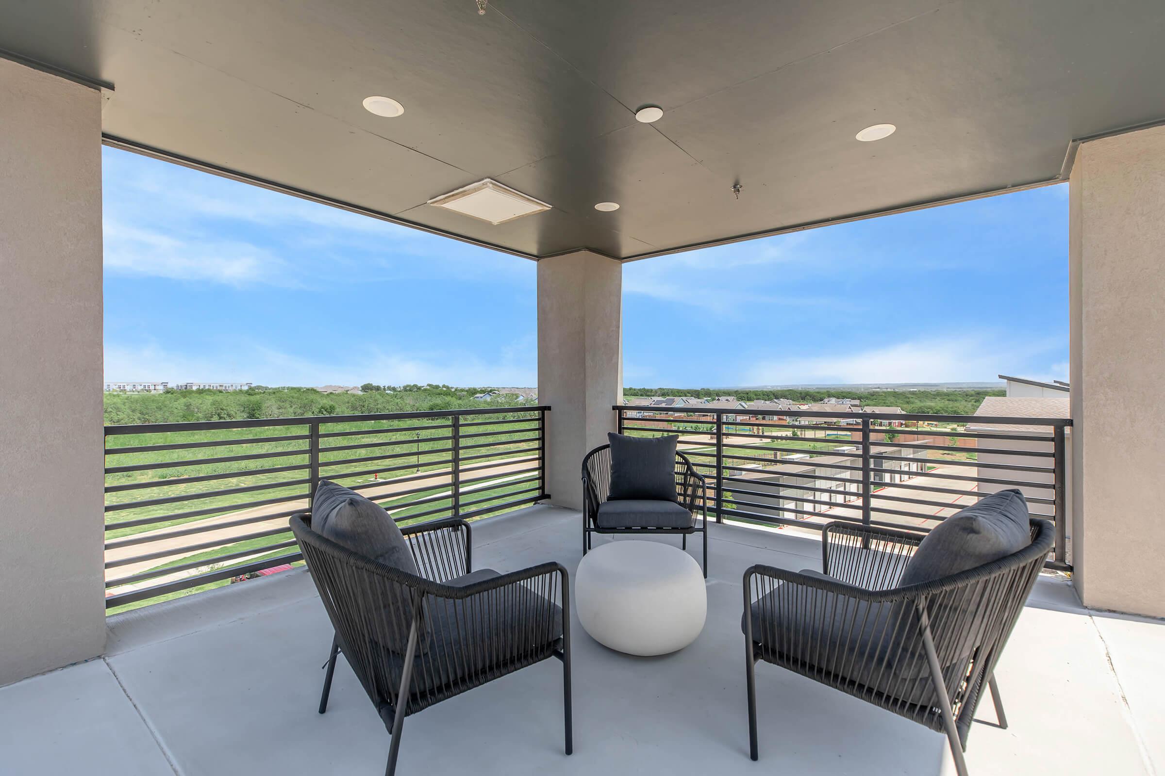 A modern outdoor balcony featuring two black wicker chairs with cushions, a round white table in the center, and a scenic view of green hills and blue sky. The space is well-lit with overhead lights, creating an inviting spot for relaxation.