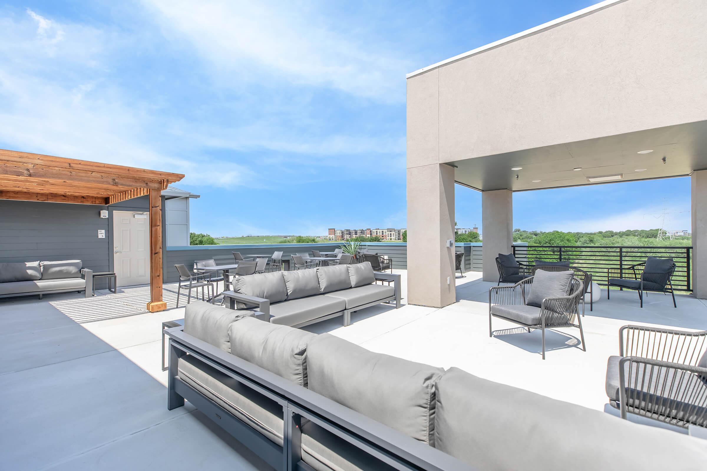Rooftop patio featuring modern lounge furniture, including gray sofas and black chairs. The space is bright and spacious, with a clear blue sky and a view of surrounding greenery and buildings. A wooden pergola adds an attractive architectural element to the area.