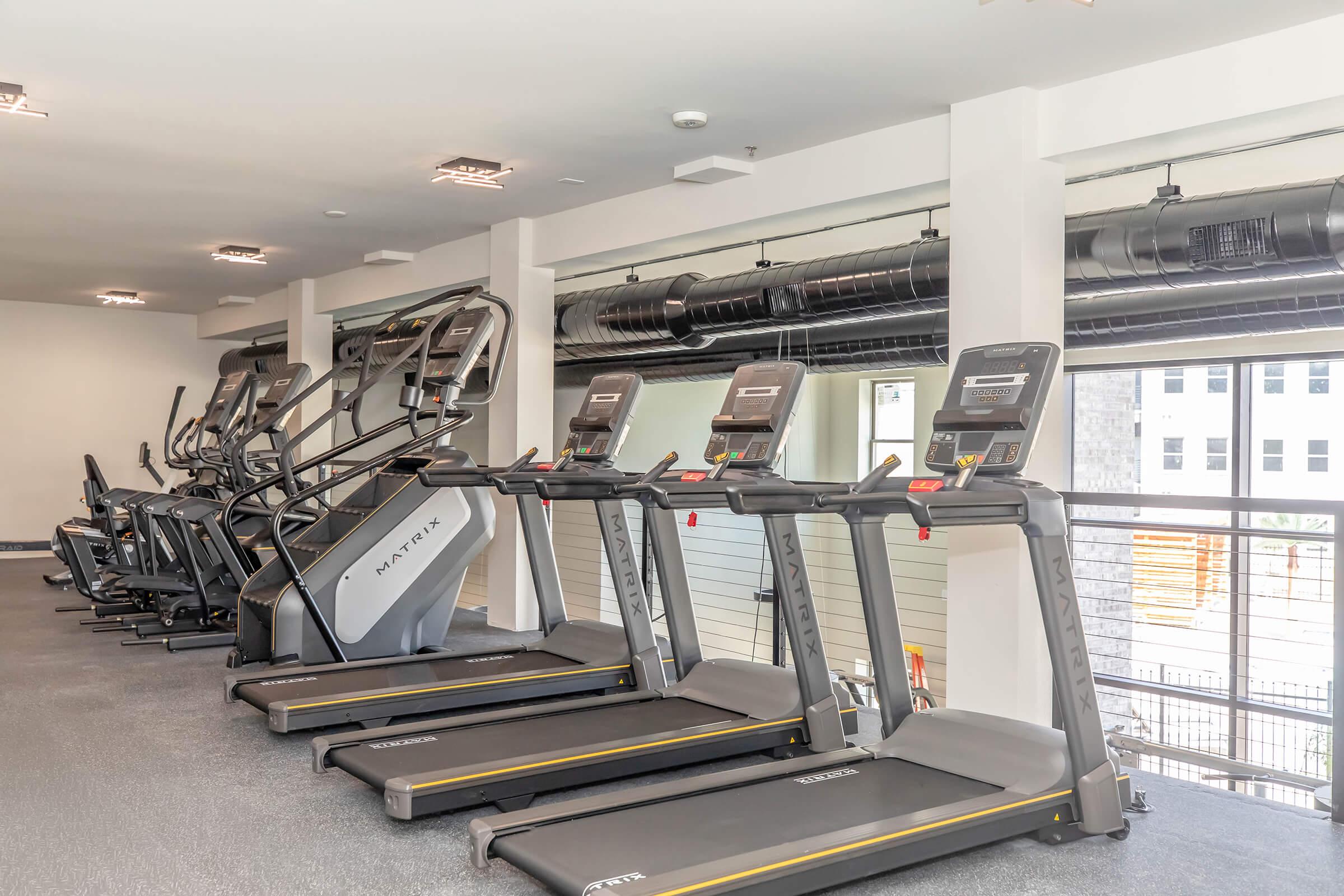 A modern gym interior featuring several treadmills and elliptical machines lined up. The space has large windows providing natural light, and the flooring is carpeted. The walls are neutral in color, and there are overhead lights illuminating the area.