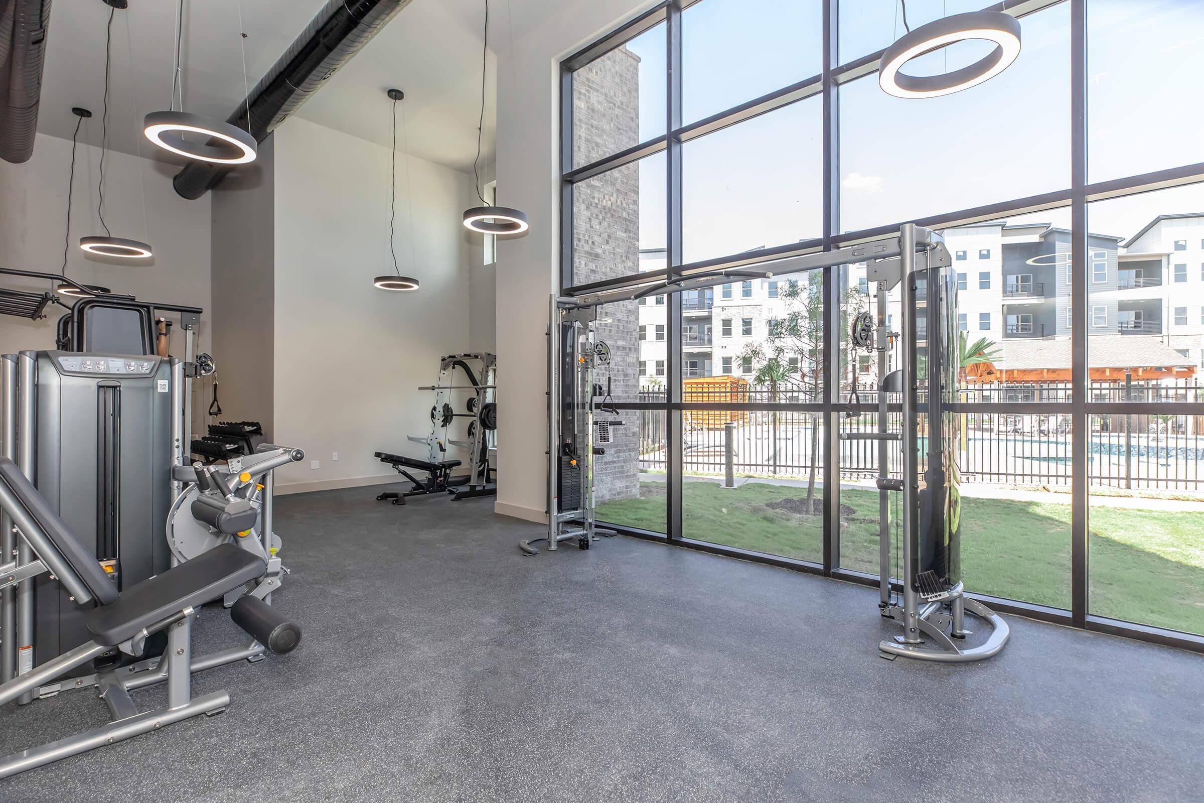 Interior view of a modern gym featuring large windows that let in natural light. The space includes various fitness equipment, such as weight machines and cable machines, against a backdrop of green grass visible through the windows. Brightly lit with contemporary lighting fixtures.