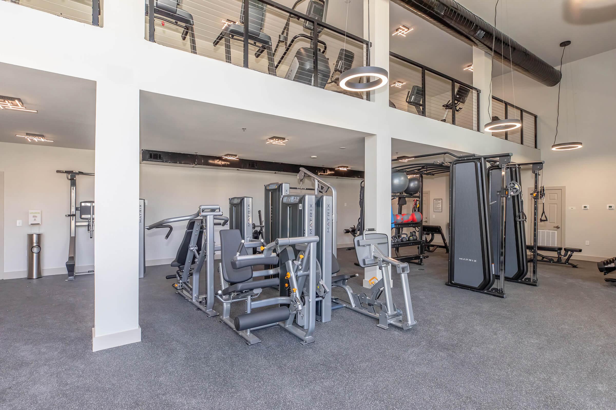 A modern gym interior featuring various exercise equipment, including weight machines and benches. The space has a clean design with gray carpeting, ample lighting, and a high ceiling. Some workout machines are positioned in the foreground, while additional equipment is visible in the background.