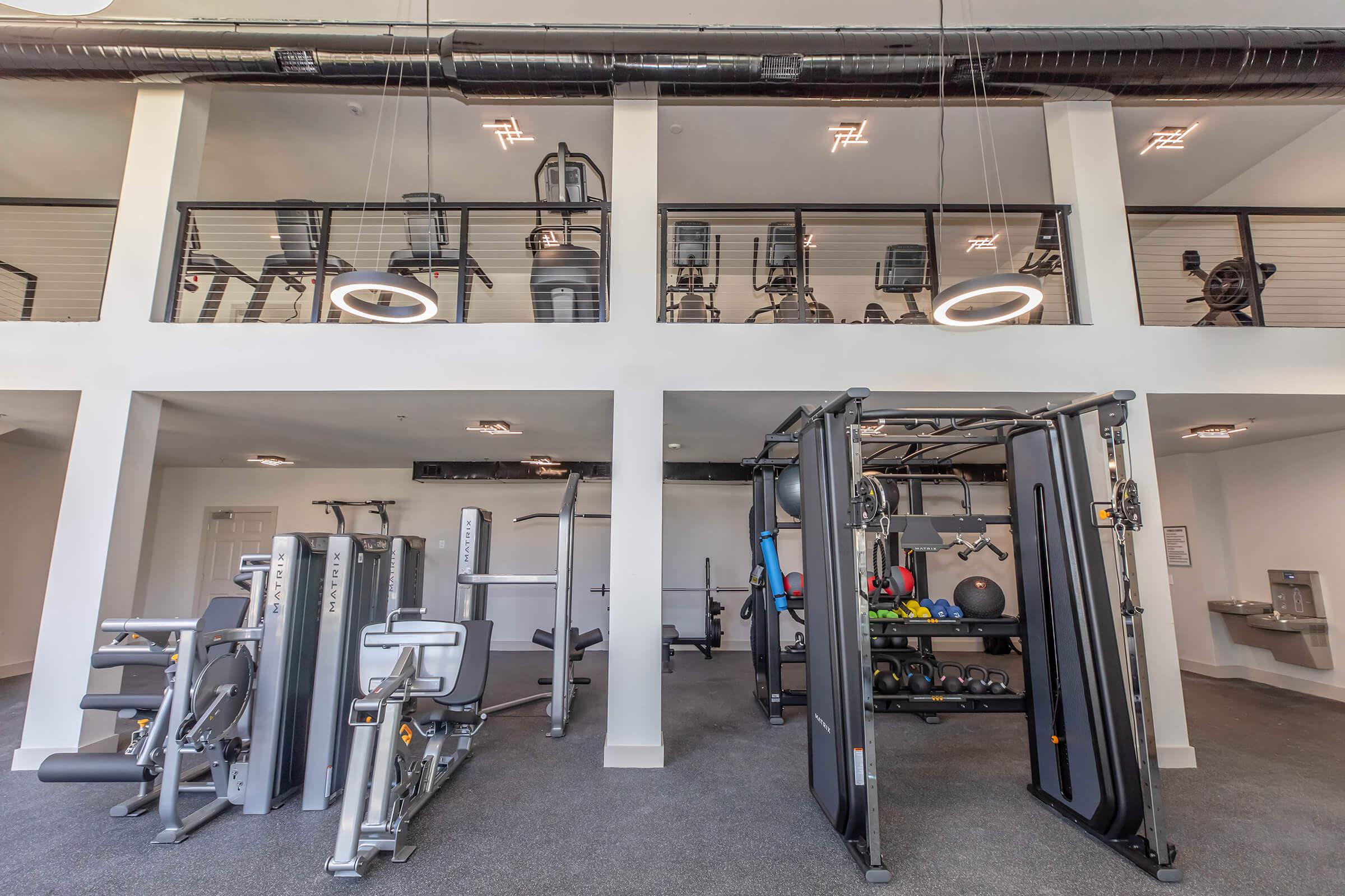 A spacious modern gym featuring a variety of exercise equipment. The lower level includes machines for strength training, weights, and benches, while the upper level houses cardio machines like stationary bikes and treadmills, all within a clean, well-lit environment.