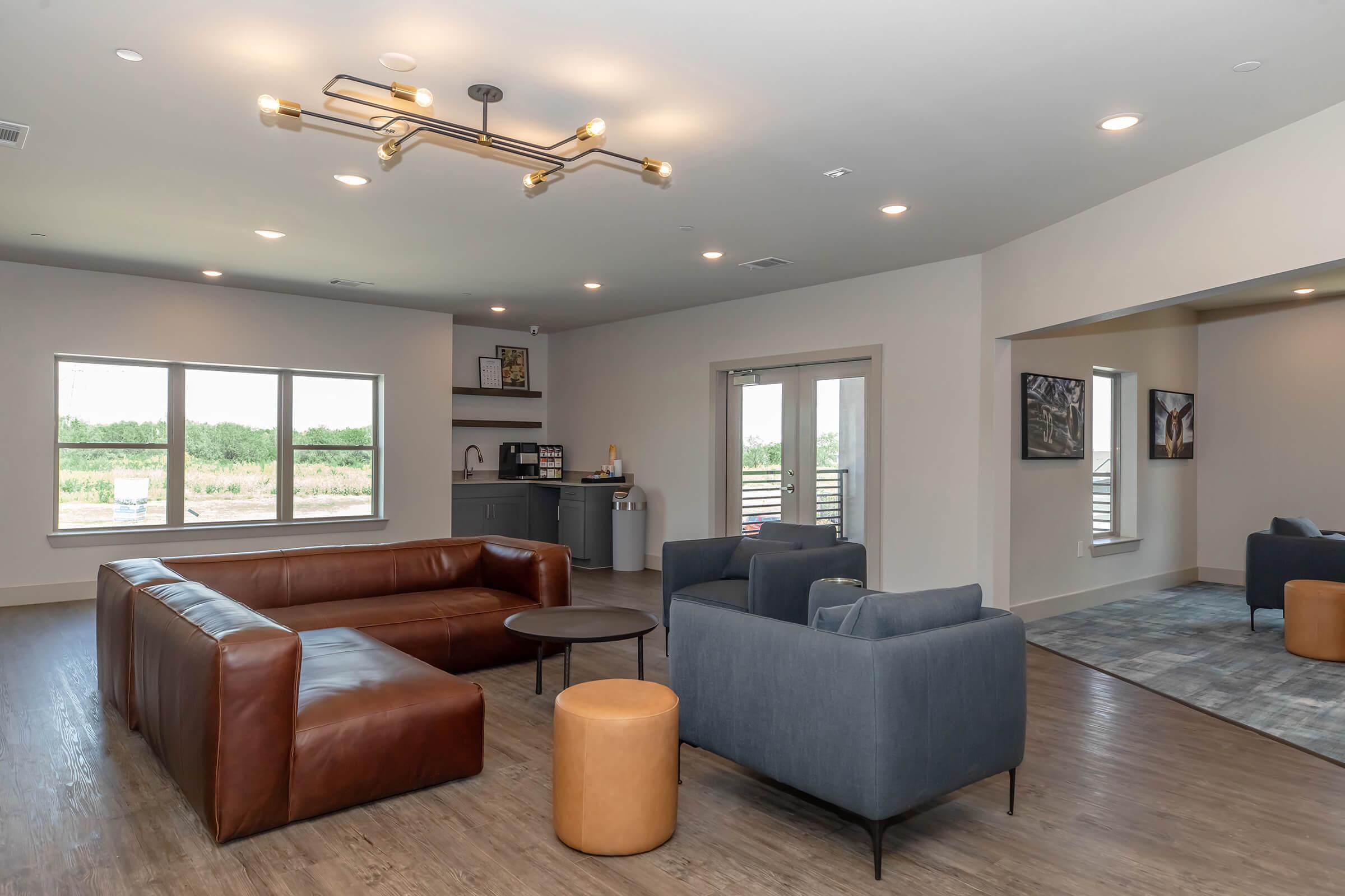 A modern lounge area featuring a brown leather sectional sofa, blue armchairs, and an orange stool. There's a round coffee table in the center, and large windows letting in natural light, showcasing a view of greenery outside. The decor is minimalist and stylish, with a neutral color palette.