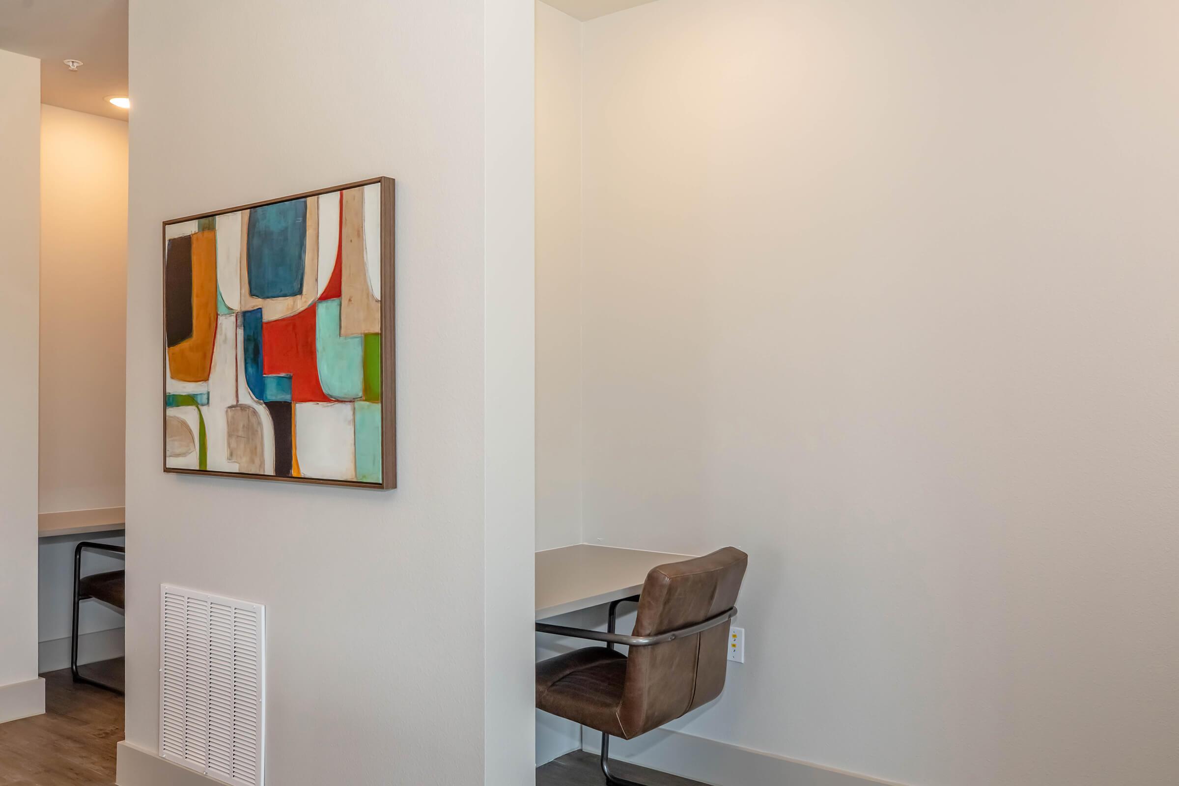 A modern interior corner featuring a colorful abstract painting on the wall. There is a small desk with a leather chair, with a clean, minimalist design. The walls are painted in a light color, contributing to an open and airy feel in the space.